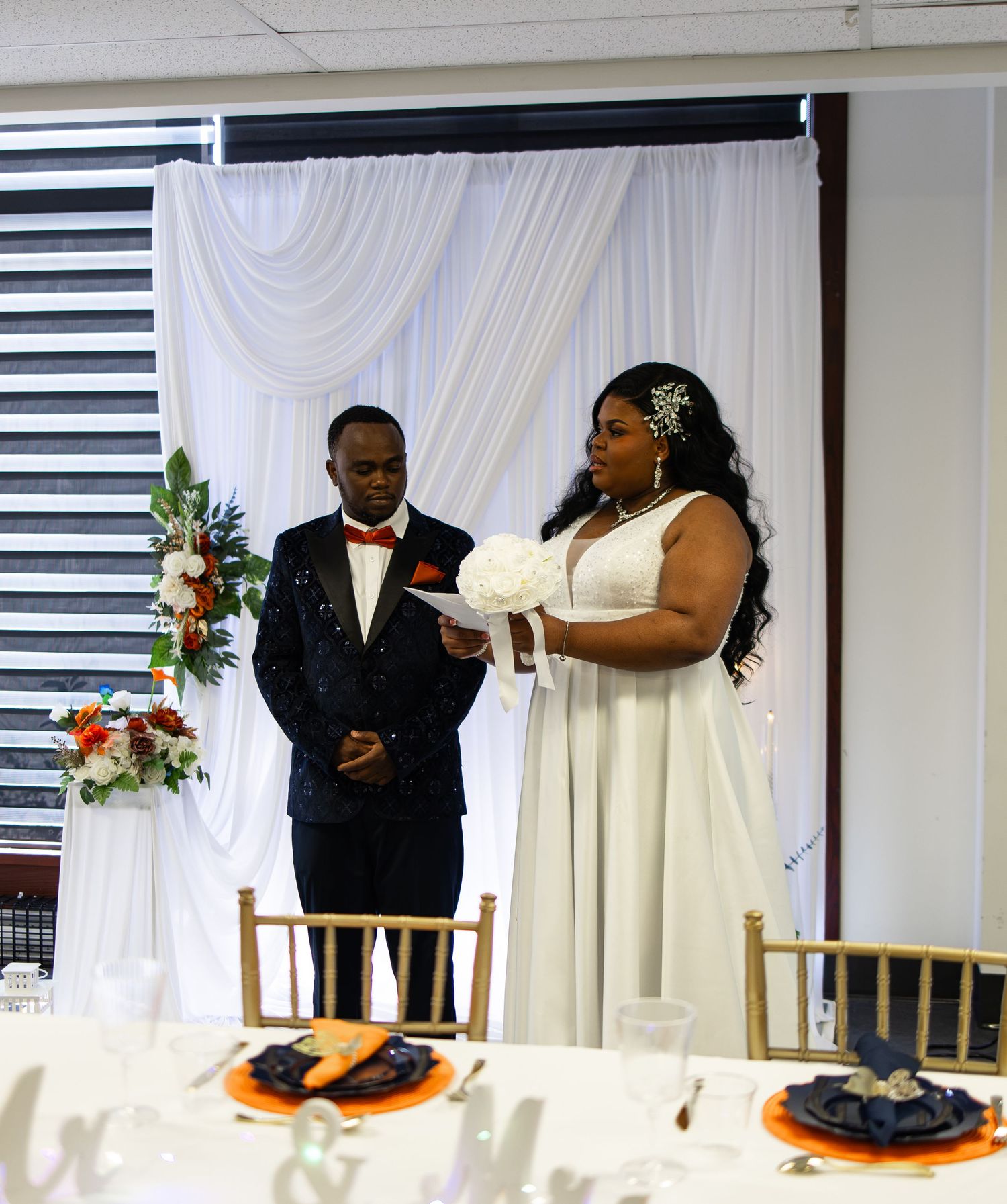 Bride in white gown holding bouquet stands with groom in tuxedo at elegant wedding ceremony with draped backdrop.