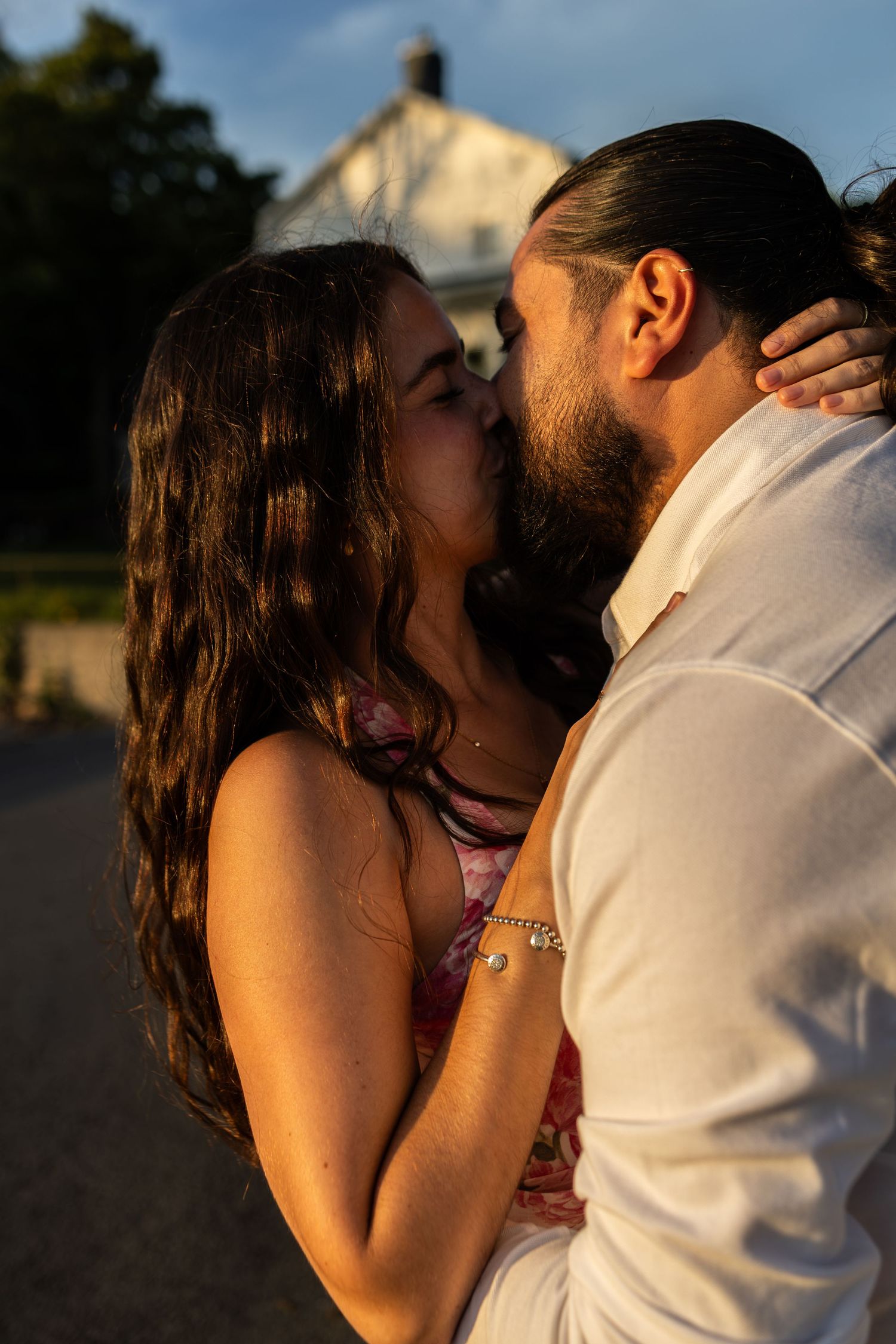 A couple shares a romantic kiss outdoors at golden hour, with a white building and trees in the background.