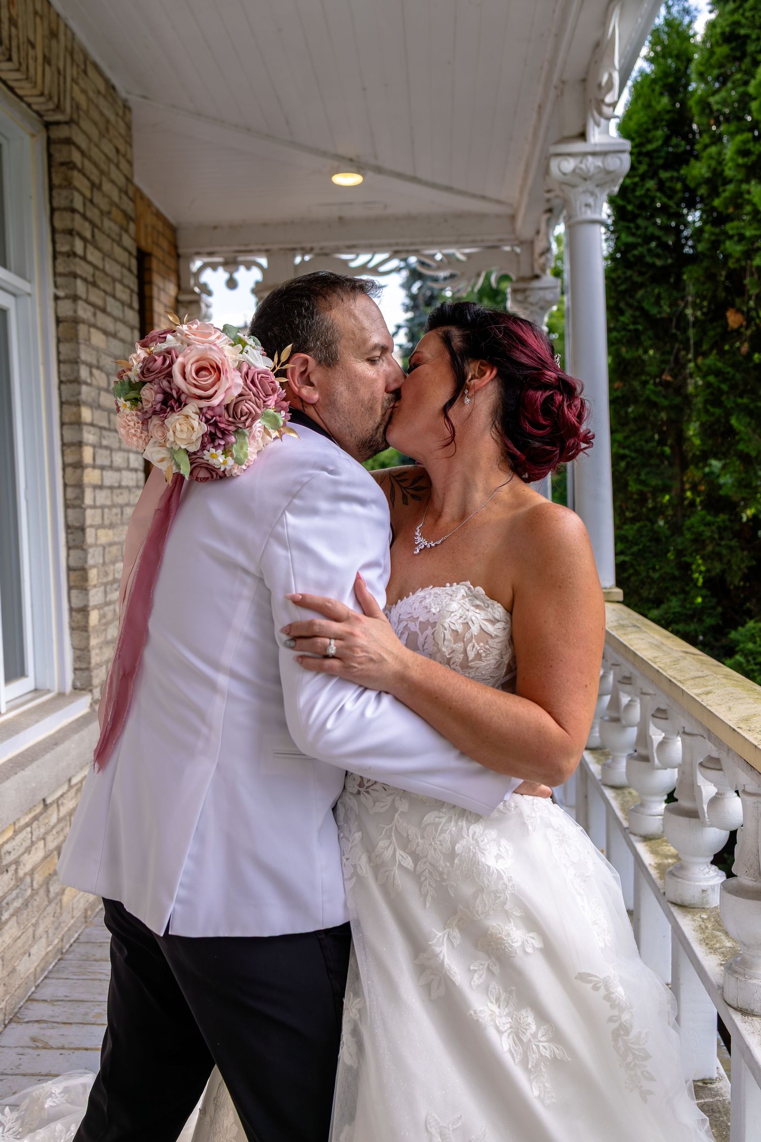 Bride and groom share a romantic kiss on a white balcony, bride holding pink bouquet in lace gown.