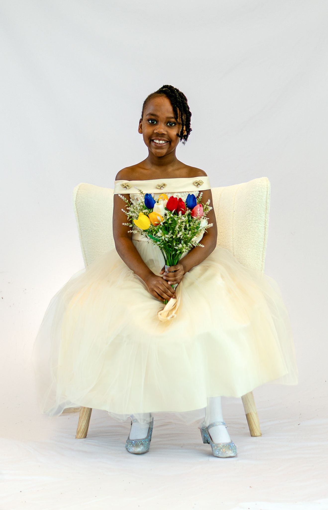 Young girl in cream off-shoulder tulle dress holds colorful flower bouquet while seated in white chair for formal portrait.