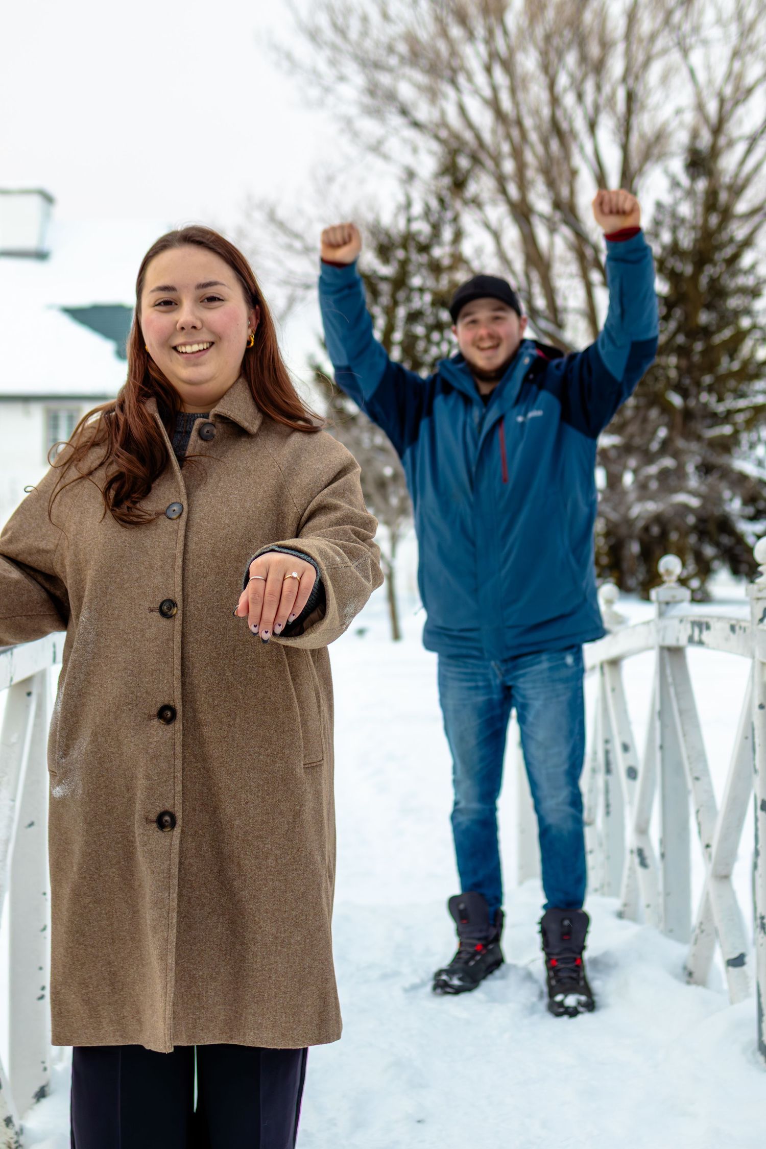 Young woman in tan coat extends hand toward camera while man in blue jacket celebrates with arms raised in snowy winter scene.