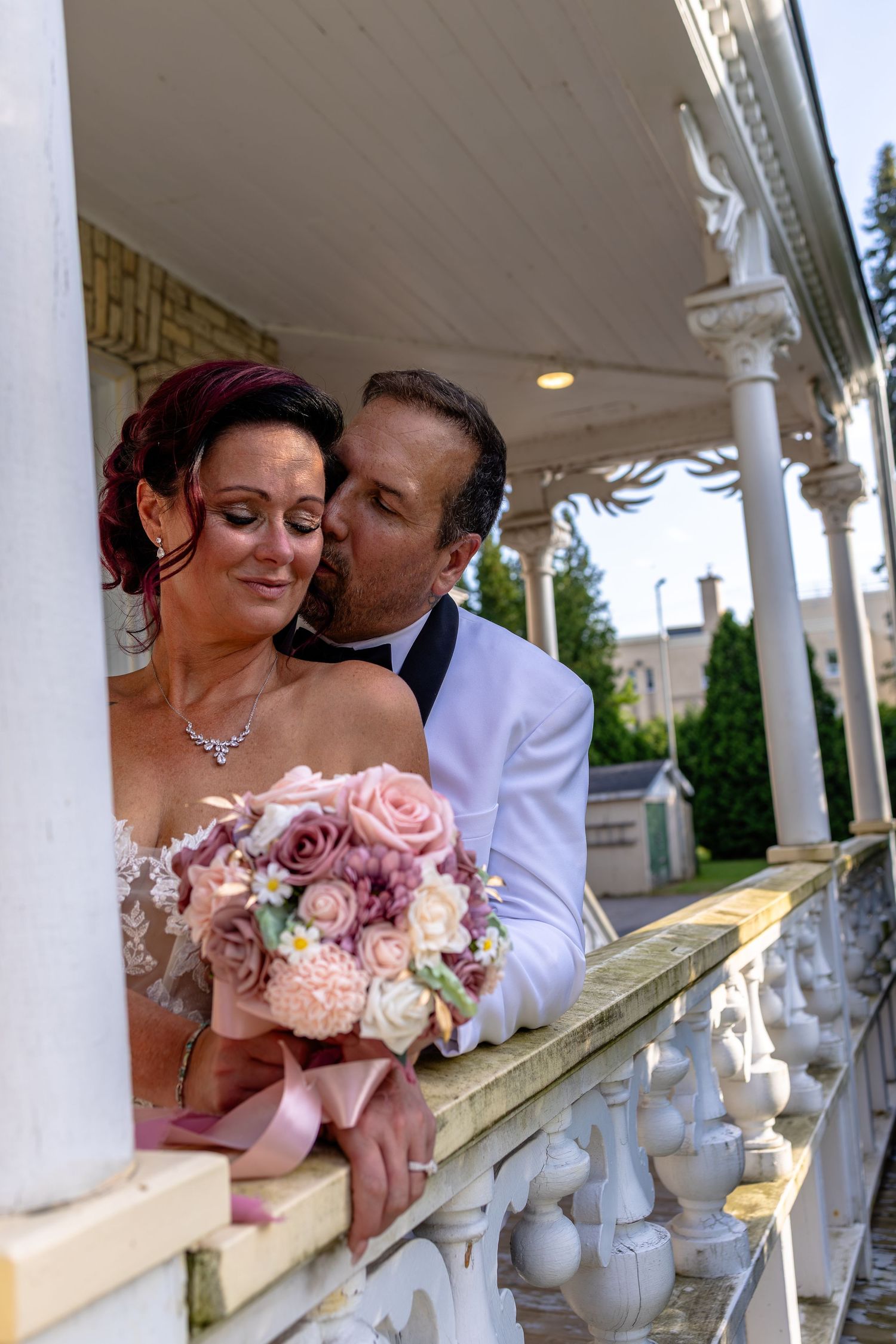Groom in white tuxedo kissing bride on cheek as she holds pink rose bouquet on elegant white balcony.