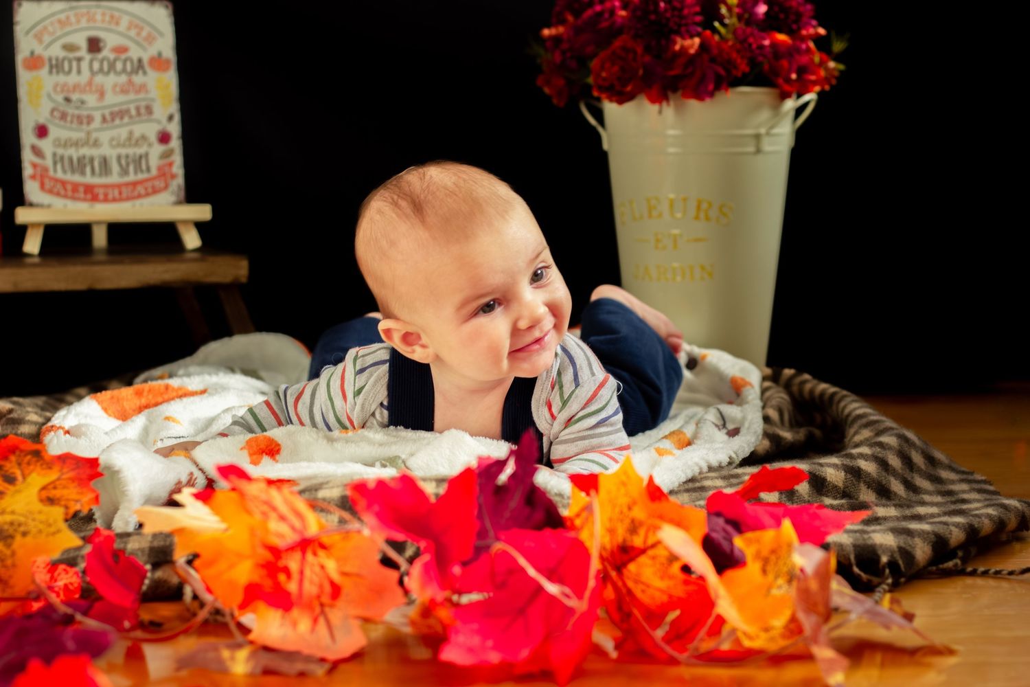 Cheerful baby doing tummy time among colorful autumn leaves with a white flower bucket nearby.