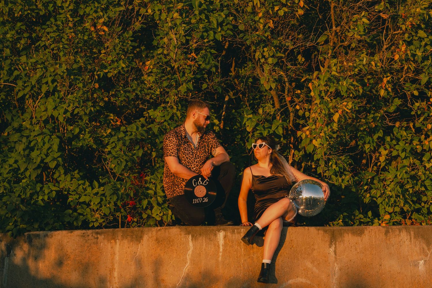 A man and woman sit on a wall surrounded by lush greenery, sharing a moment with a disco ball at golden hour.