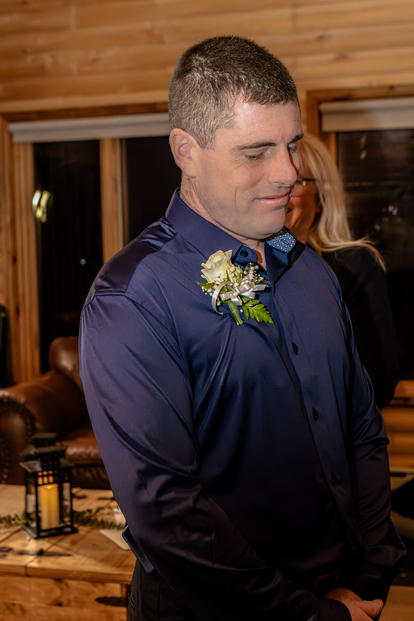 Groom in a navy blue dress shirt with a white rose boutonniere standing in a rustic log cabin setting.