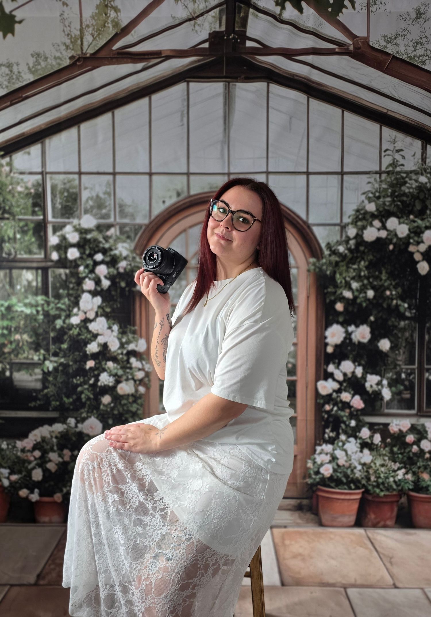 Woman in white lace dress holding camera sits before floral greenhouse backdrop.
