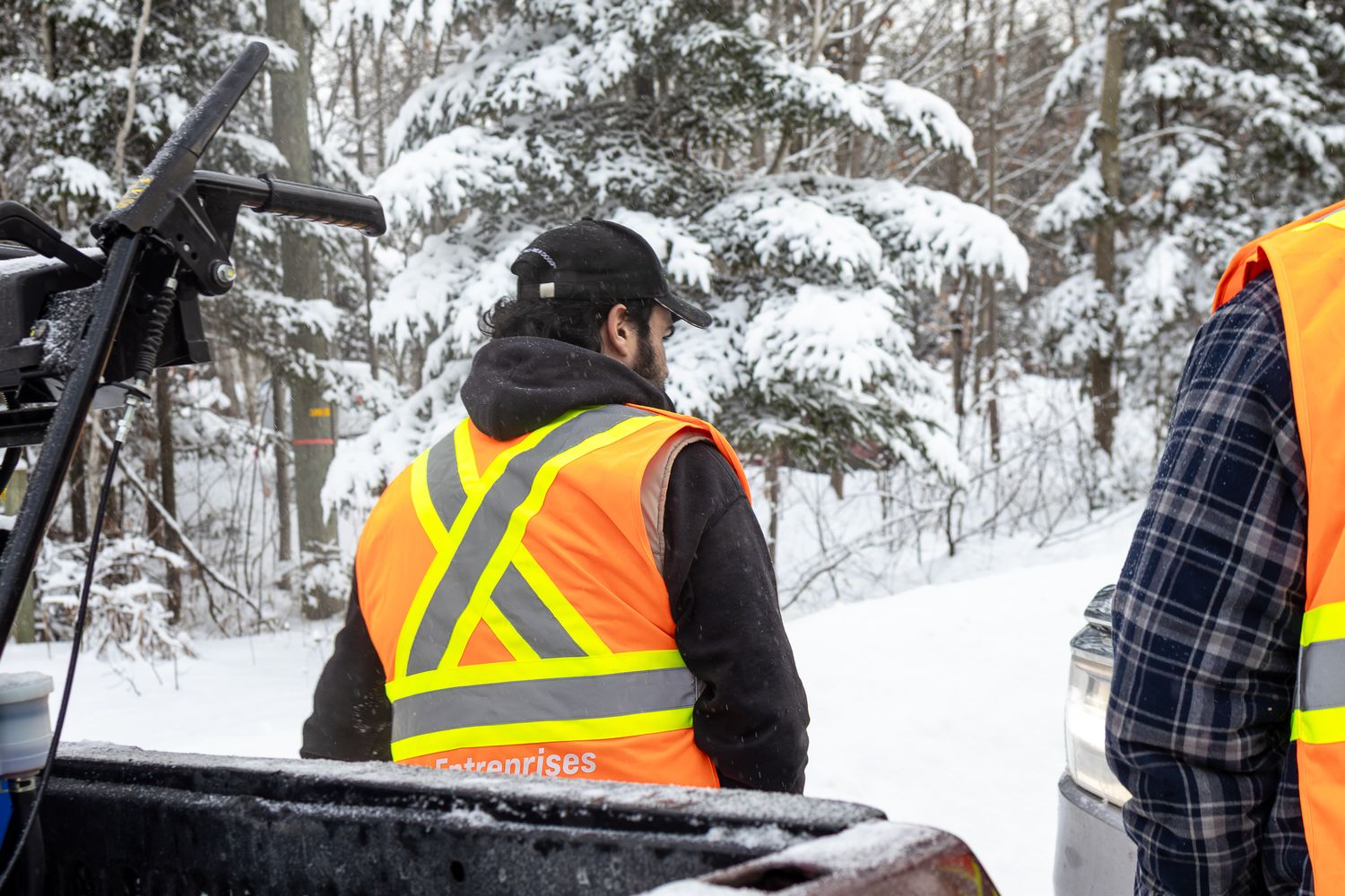 Worker in orange safety vest riding in a truck bed on a snow-covered forest trail.