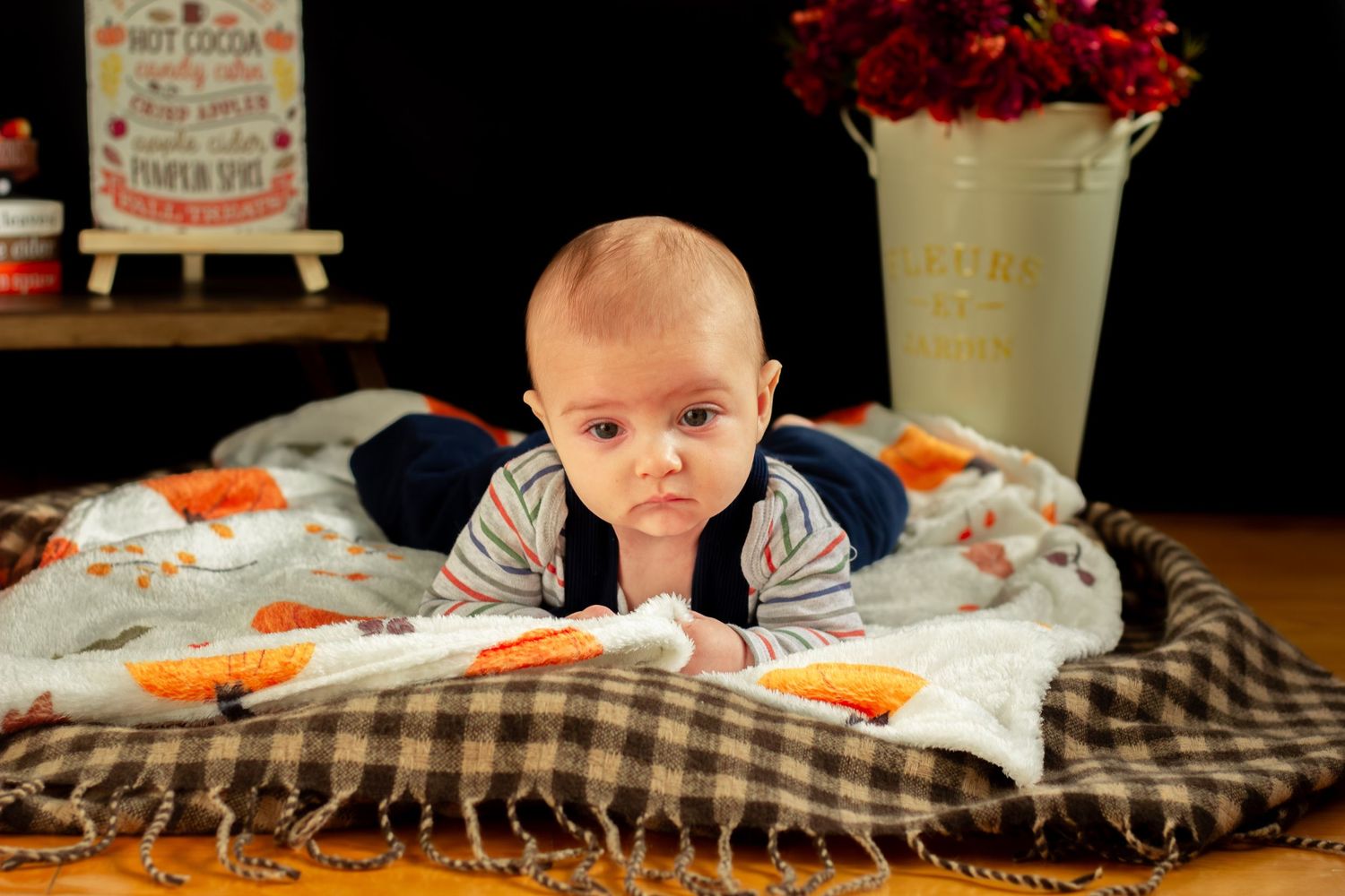 Infant in striped shirt and blue overalls during tummy time on a patterned fall blanket.