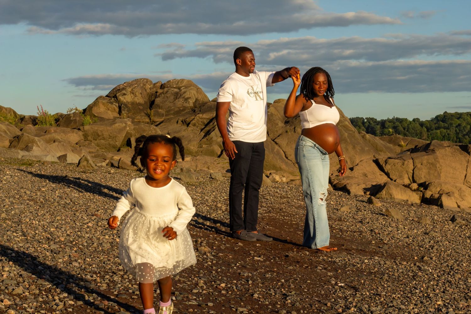 A family with a young girl in a white dress and pregnant mother poses on a rocky beach at golden hour.