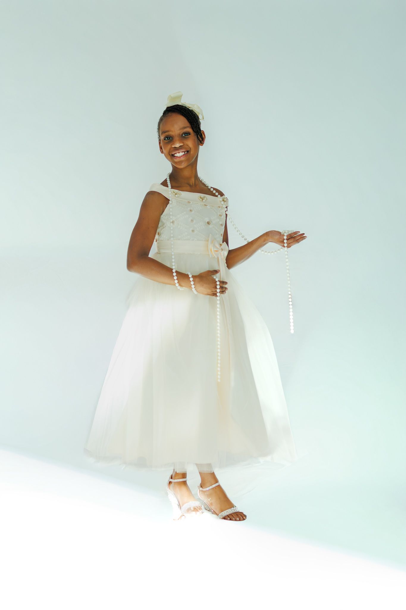 Joyful young girl in white communion dress holding pearl rosary beads on a soft blue background.