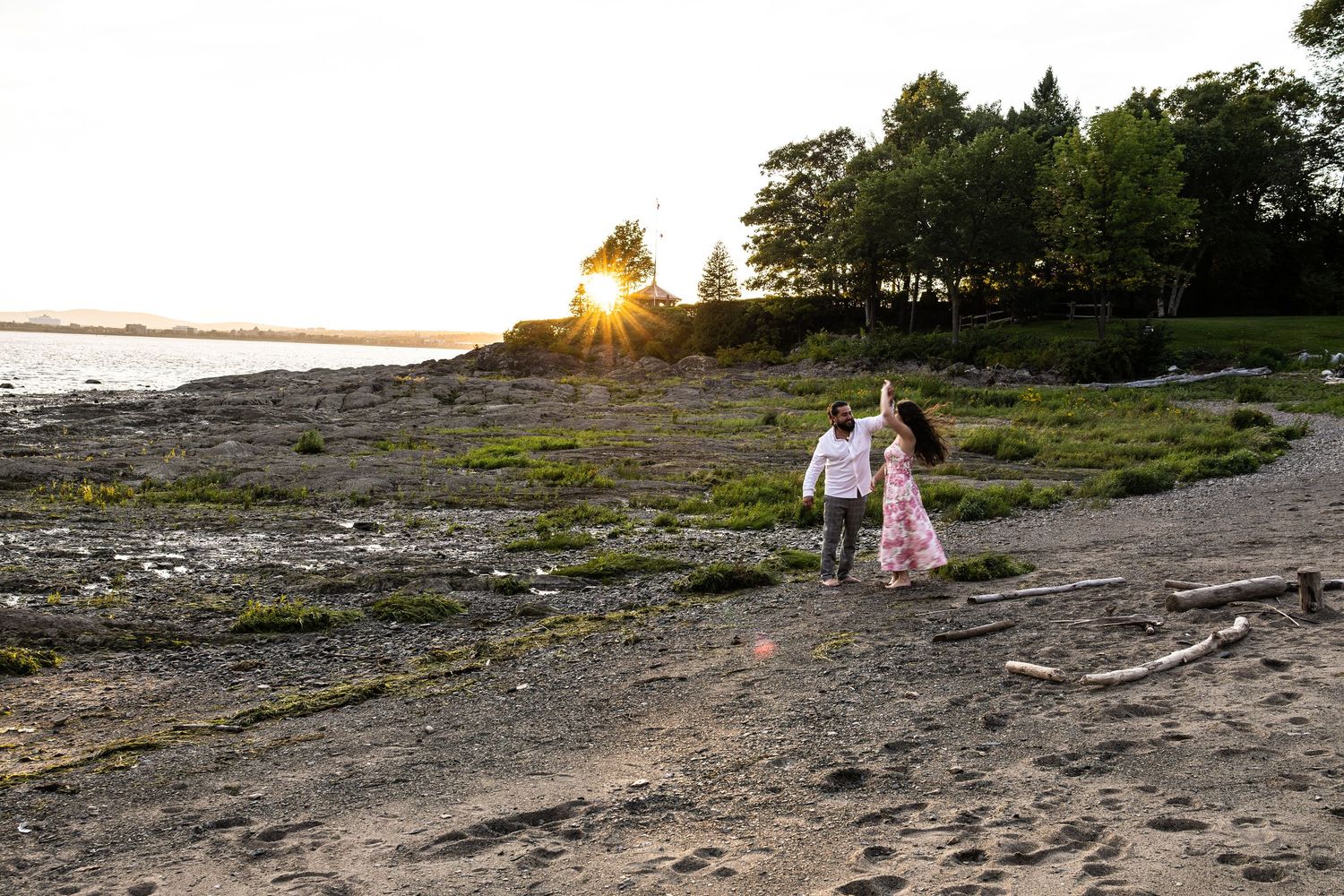 Couple walks hand-in-hand on a rocky beach at sunset with golden sunburst through trees.