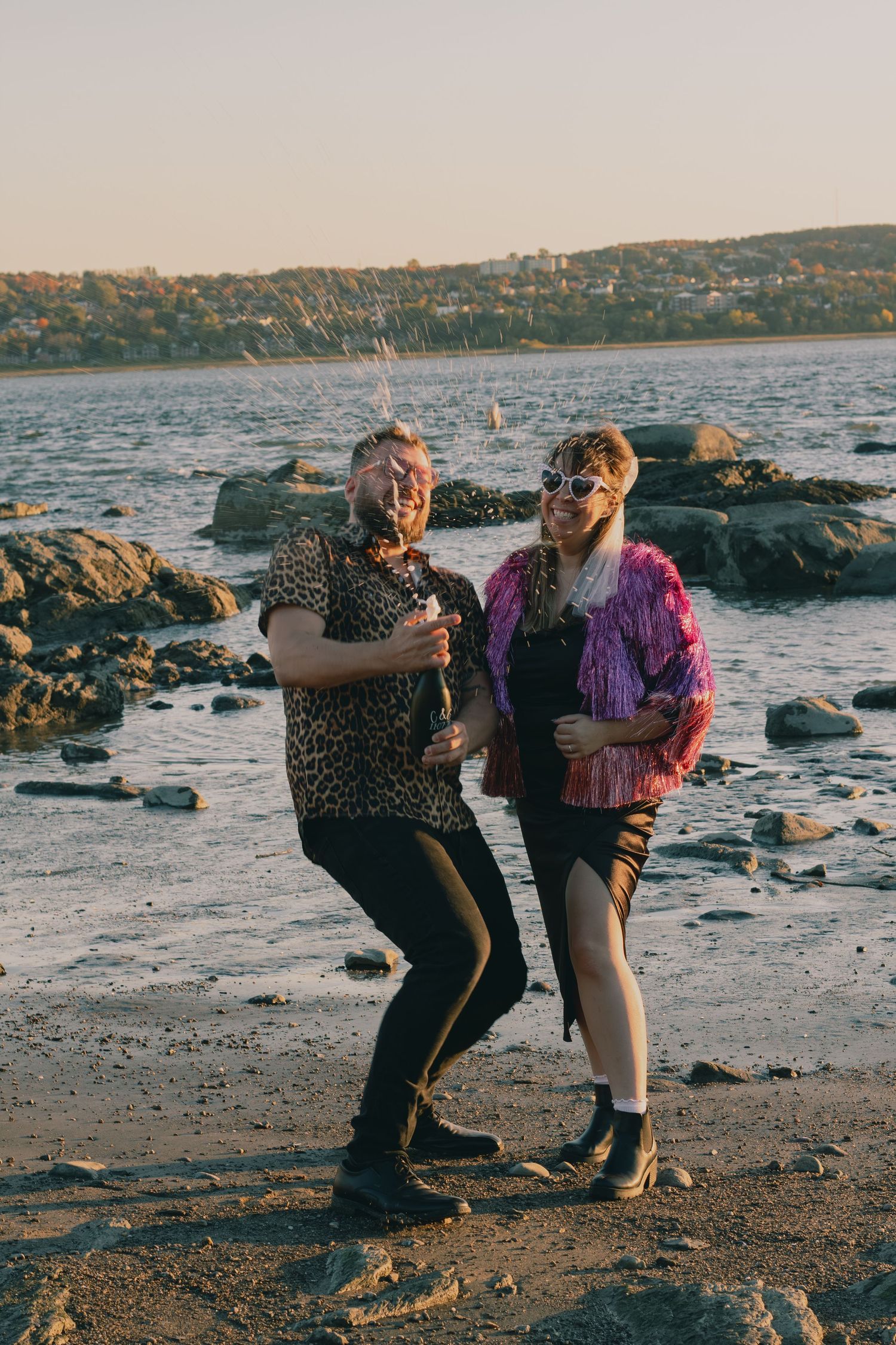 Two friends celebrate on a rocky beach at sunset, popping champagne in colorful festive outfits.