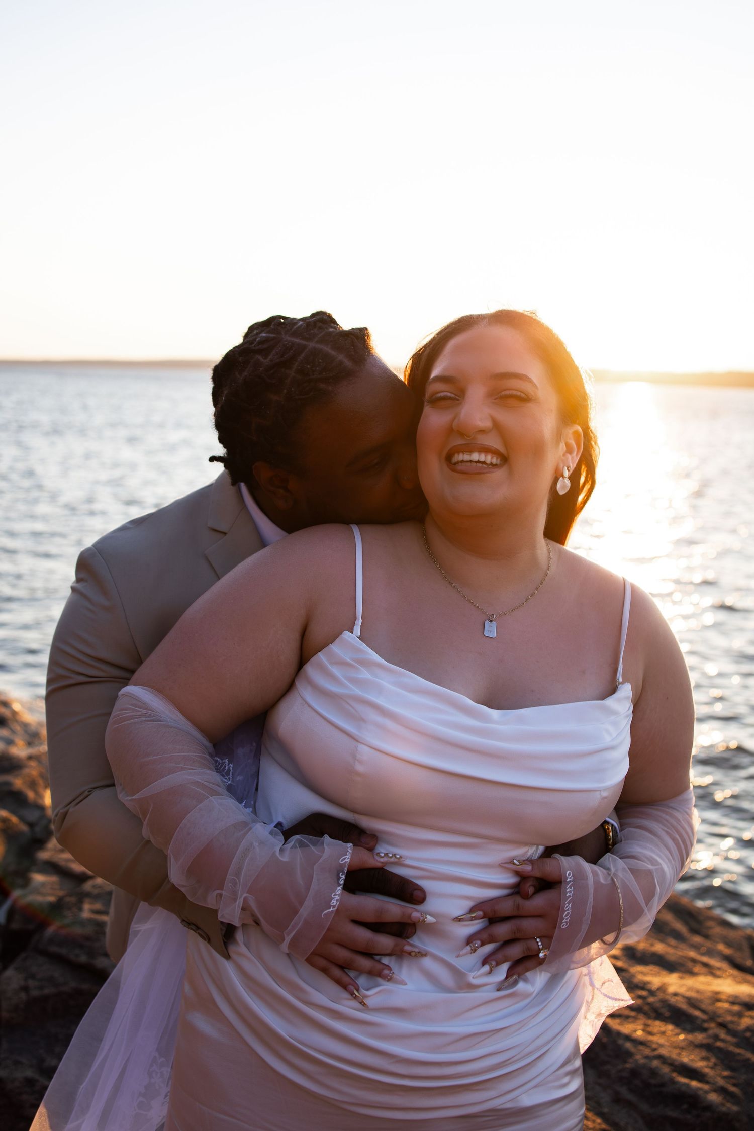 Couple in wedding attire embracing at sunset by the water, bride in white dress laughing joyfully.