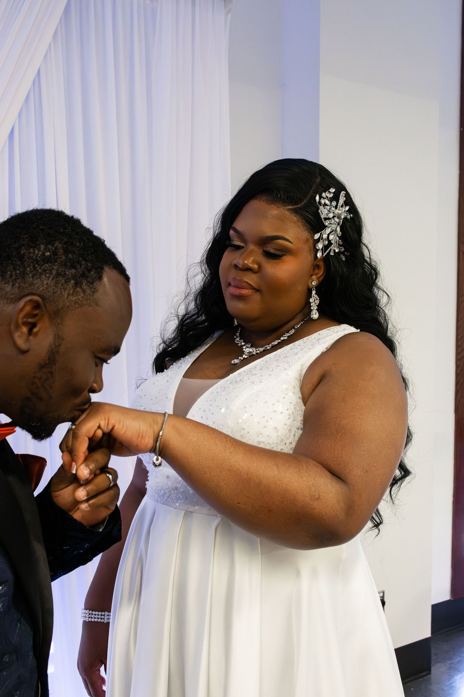 A groom kissing the hand of his smiling bride in a white wedding gown with silver hair accessories.
