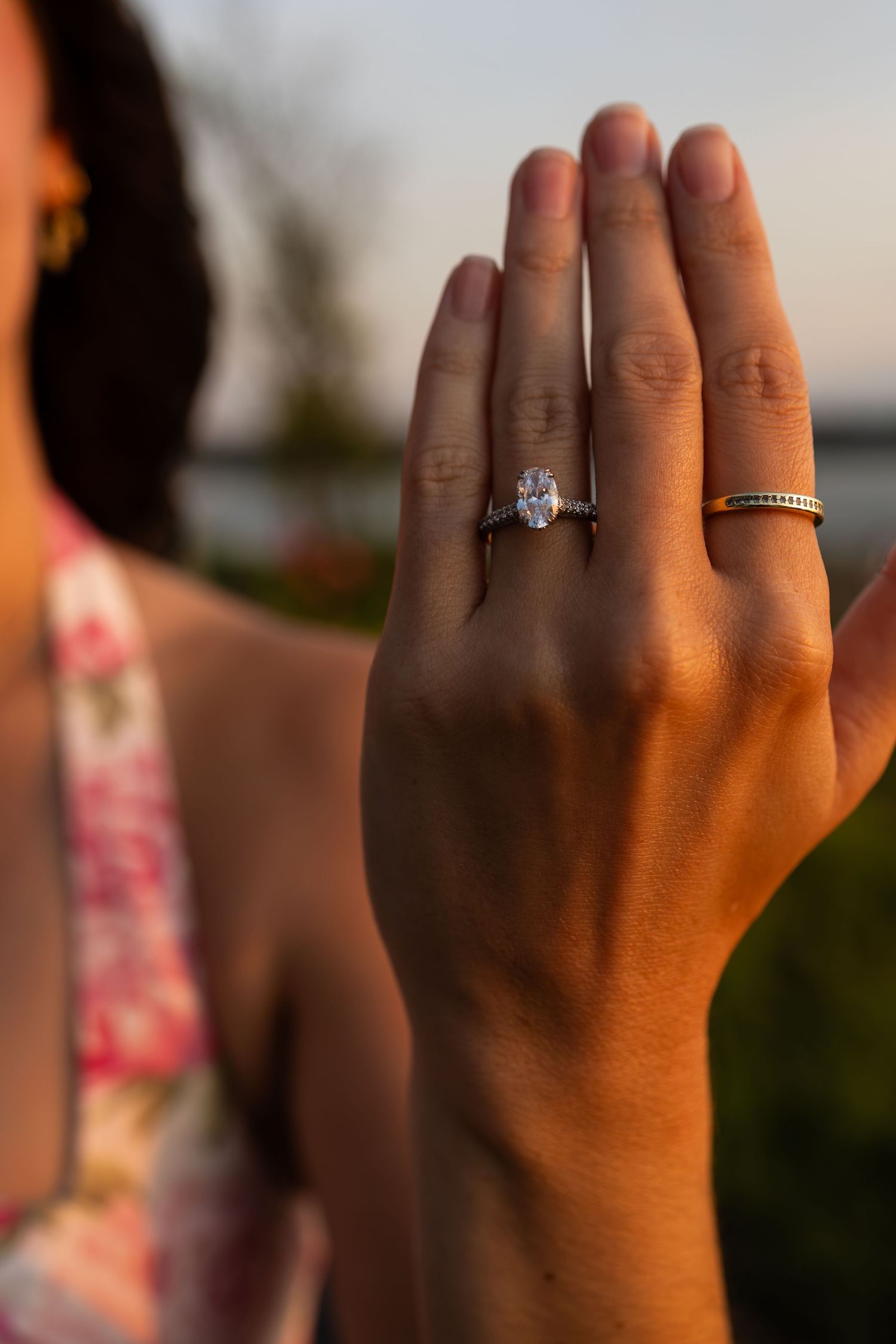 Close-up of a woman's hand displaying a stunning oval diamond engagement ring and gold band at sunset.