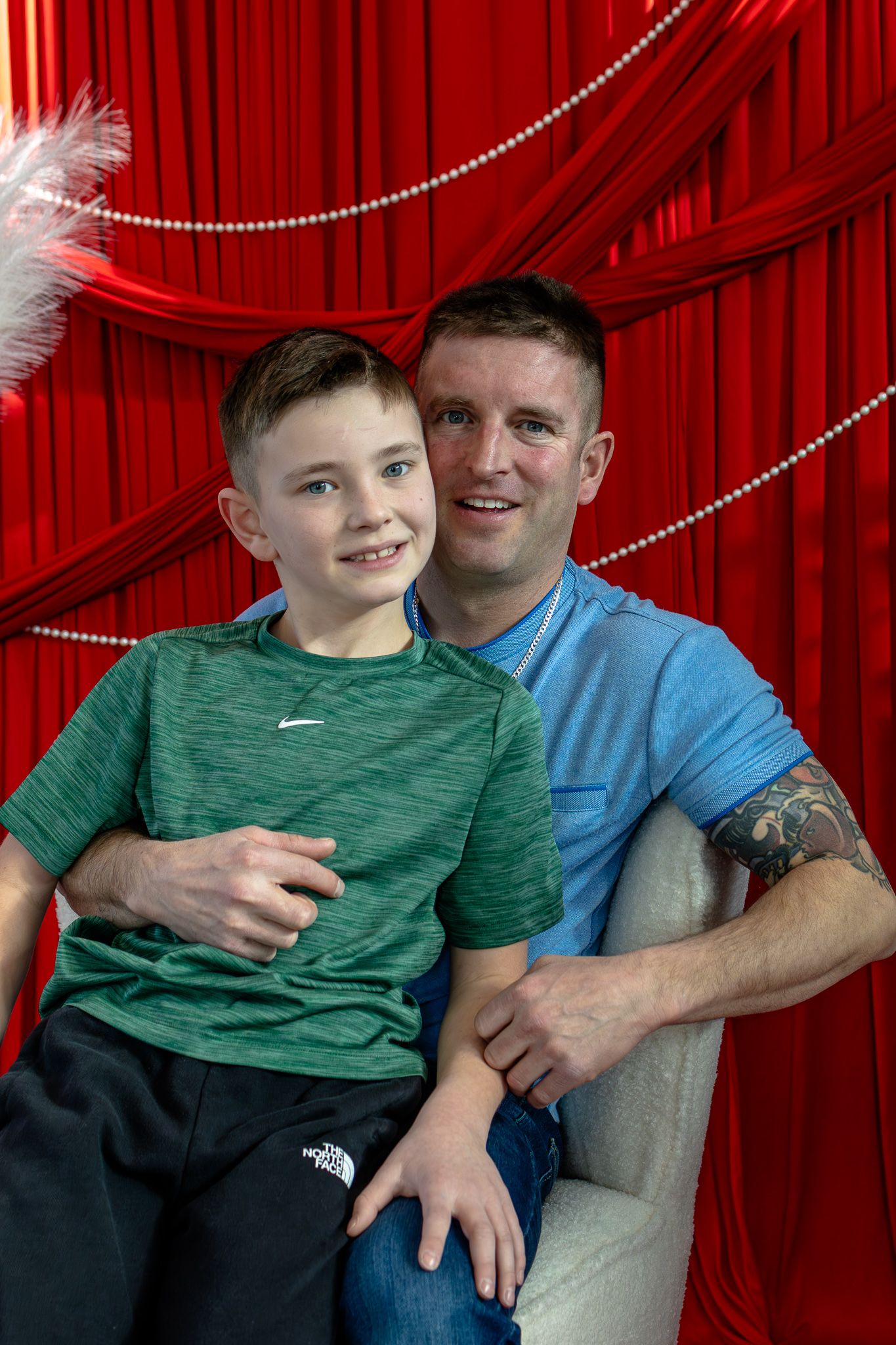 Father and preteen son posing together on a white chair against a red curtain background.