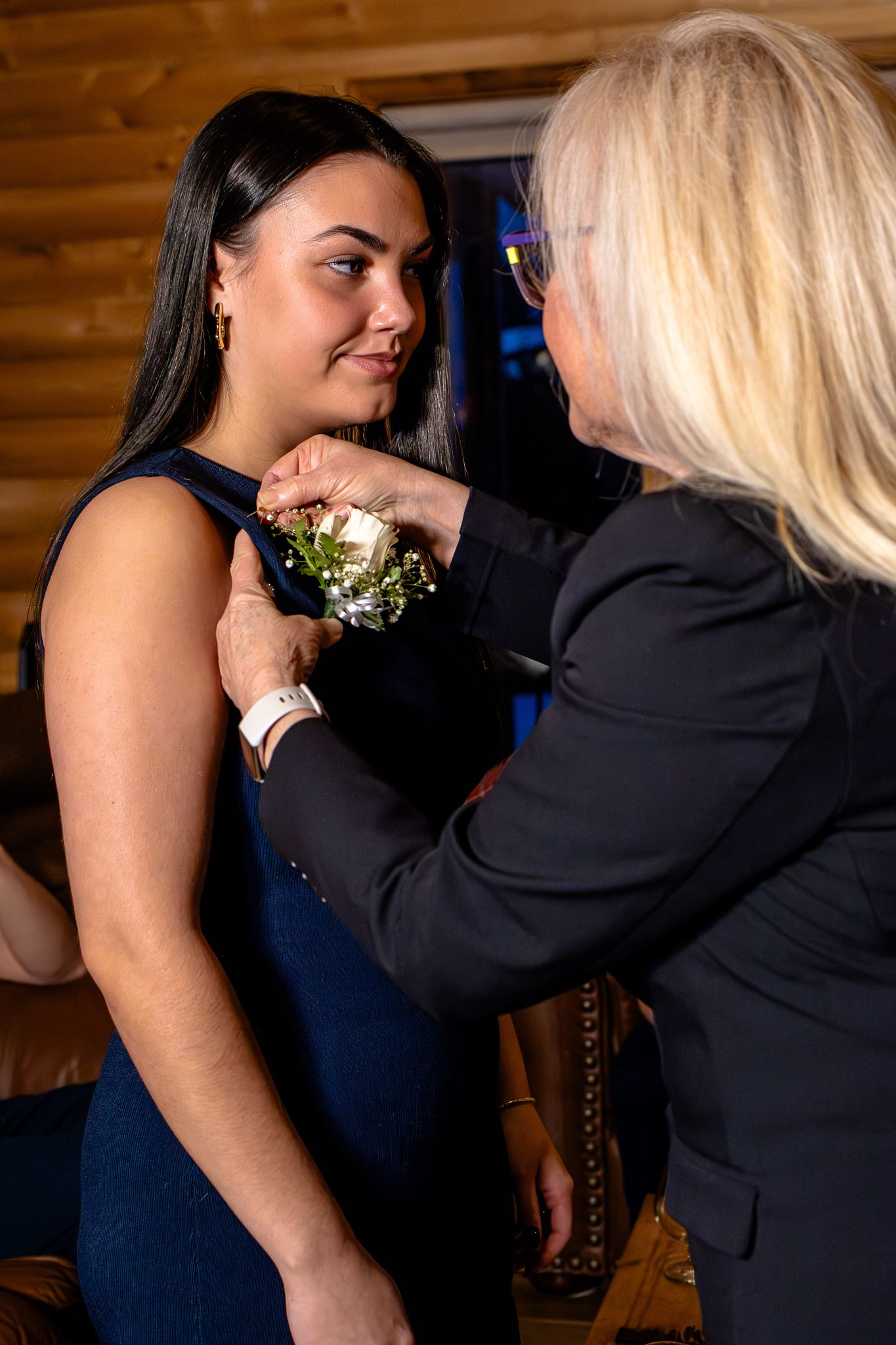 Older blonde woman pinning a white floral boutonniere on a young dark-haired woman in a navy dress.