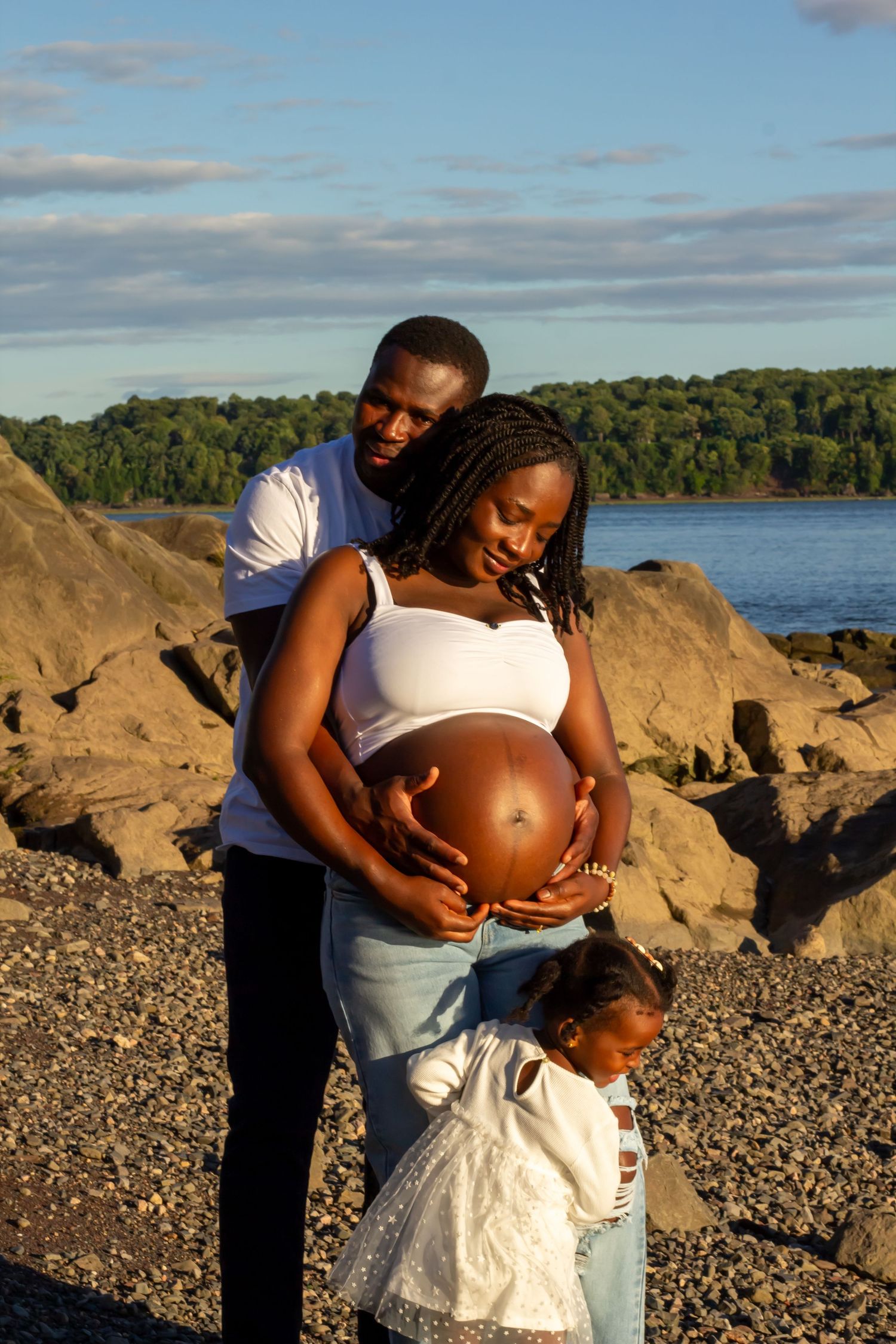 Black family with father, pregnant mother, and toddler daughter bonding on pebble beach.