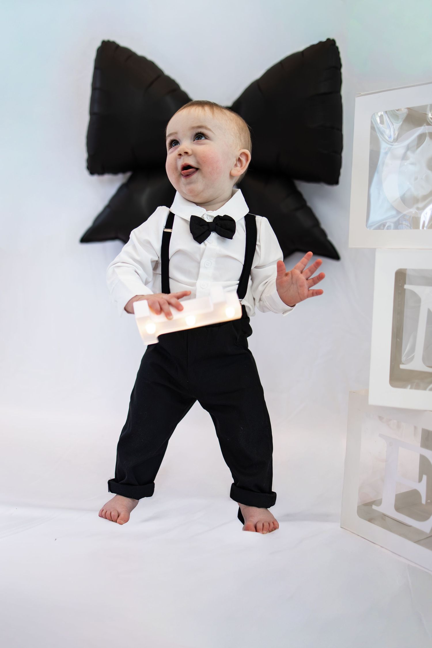 Adorable baby boy in white shirt, black bow tie, suspenders, and pants, holding a glowing gift box.
