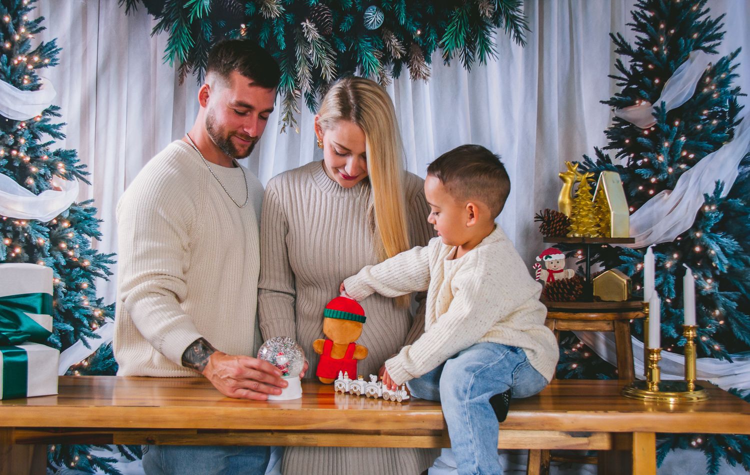 Family with young boy playing with toys at a decorated Christmas table with festive tree backdrop.