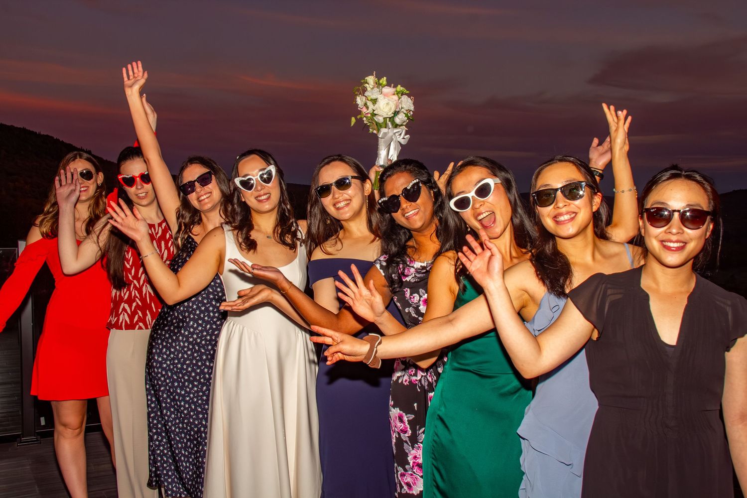 Bride and bridesmaids in sunglasses joyfully toss the wedding bouquet outdoors during a stunning sunset celebration.