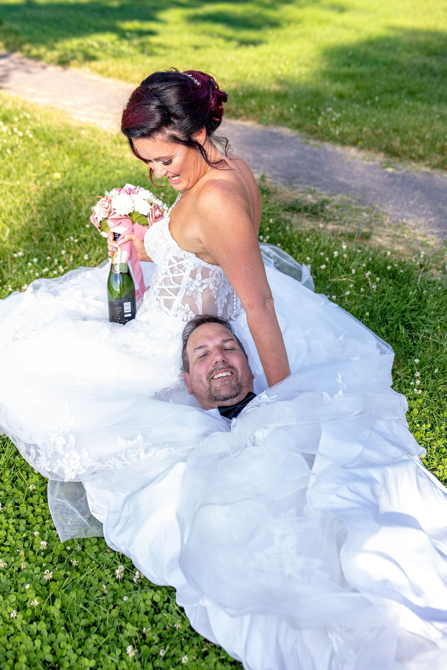 Bride in white gown sits on grass holding bouquet and champagne, with groom lying beneath her dress, smiling outdoors.