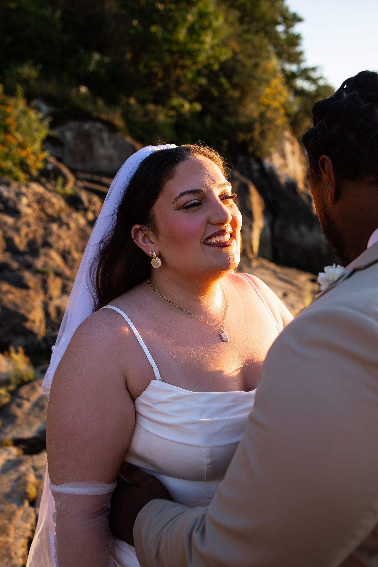 Bride in white gown and veil smiling joyfully at groom during outdoor wedding ceremony on rocky coastline at sunset.