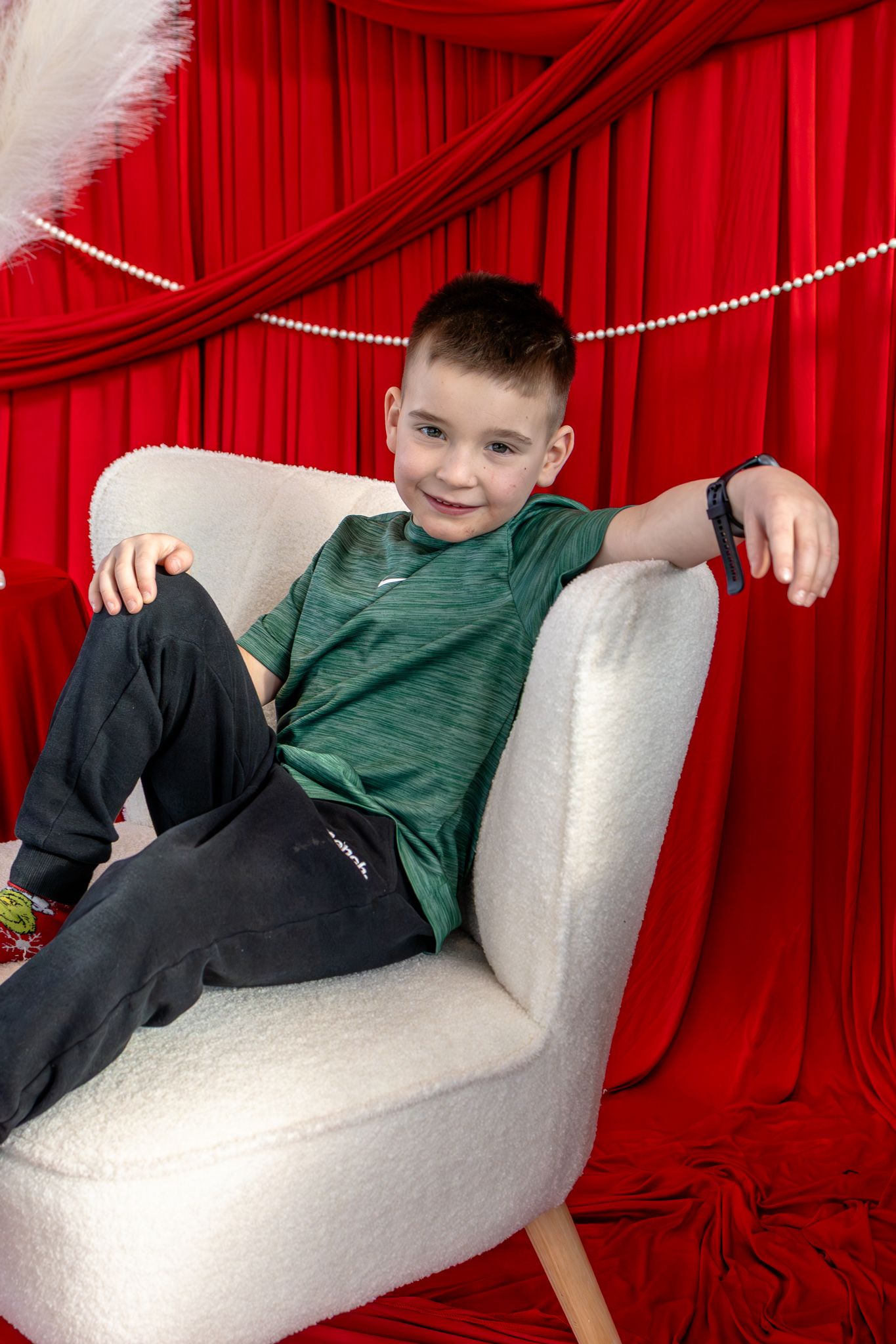Smiling boy relaxing casually in white armchair against bright red curtain studio backdrop.