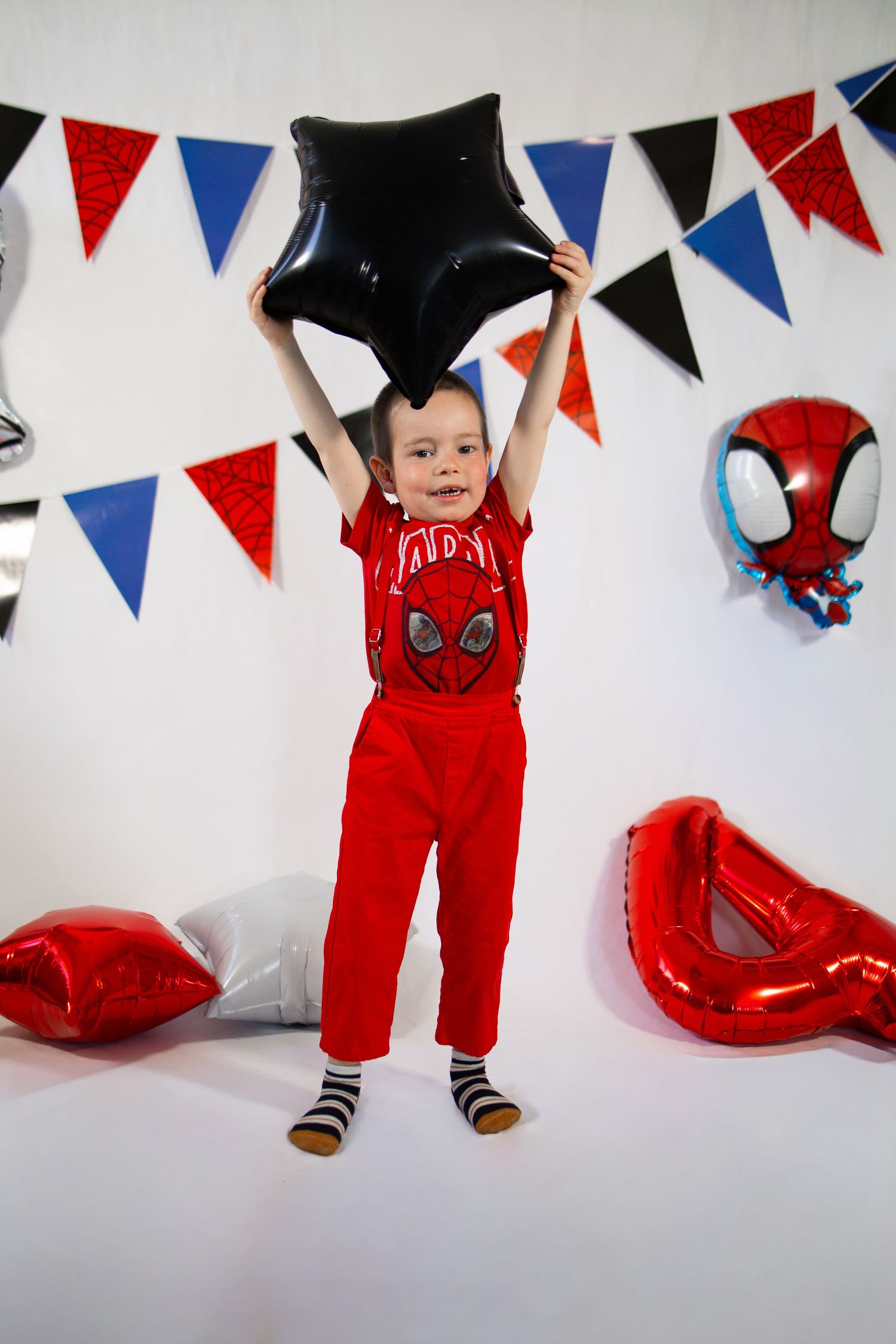 Boy in red Deadpool outfit holds black star balloon at Spiderman birthday party with colorful bunting.