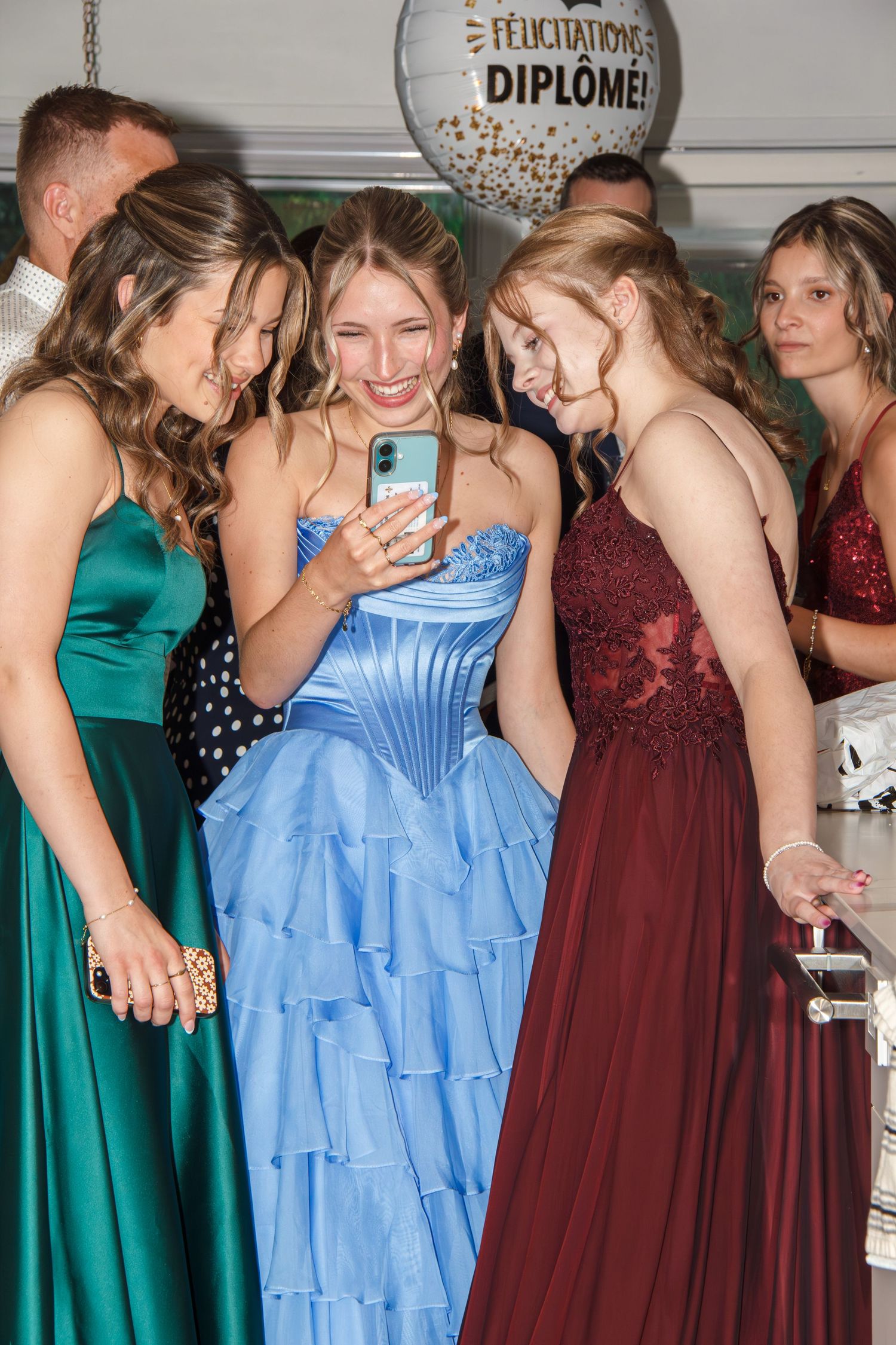 Young women in elegant prom gowns gather around a smartphone at a graduation celebration with a Felicitations Diplome balloon.