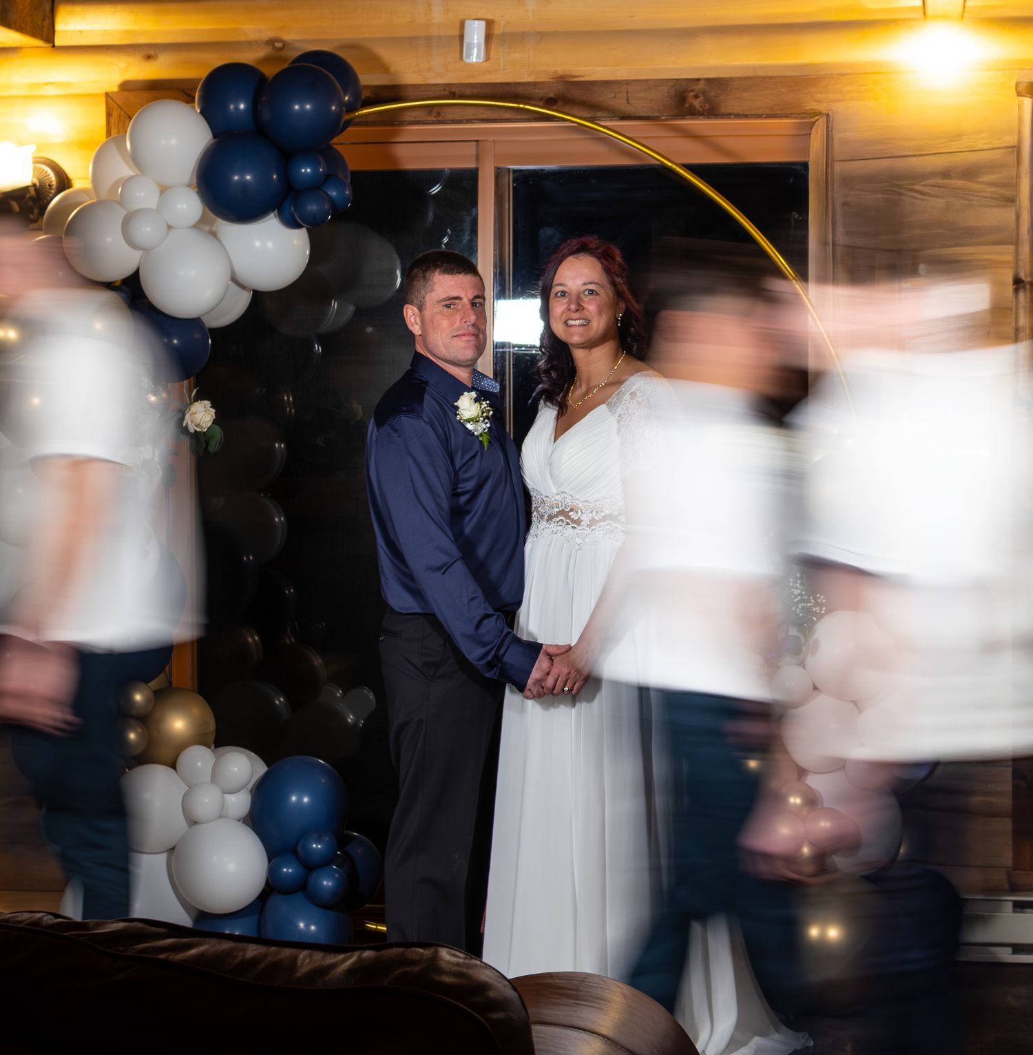 Bride and groom holding hands under golden hoop arch with navy blue and white balloon decorations.