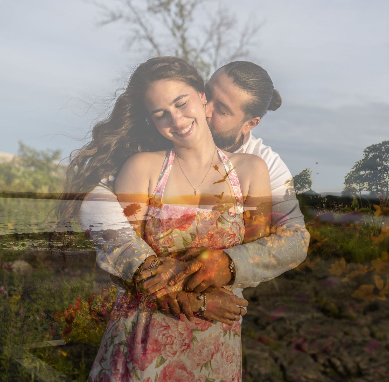 A couple embraces in a dreamy double-exposure photo, blending with a lush outdoor landscape of trees and fields.