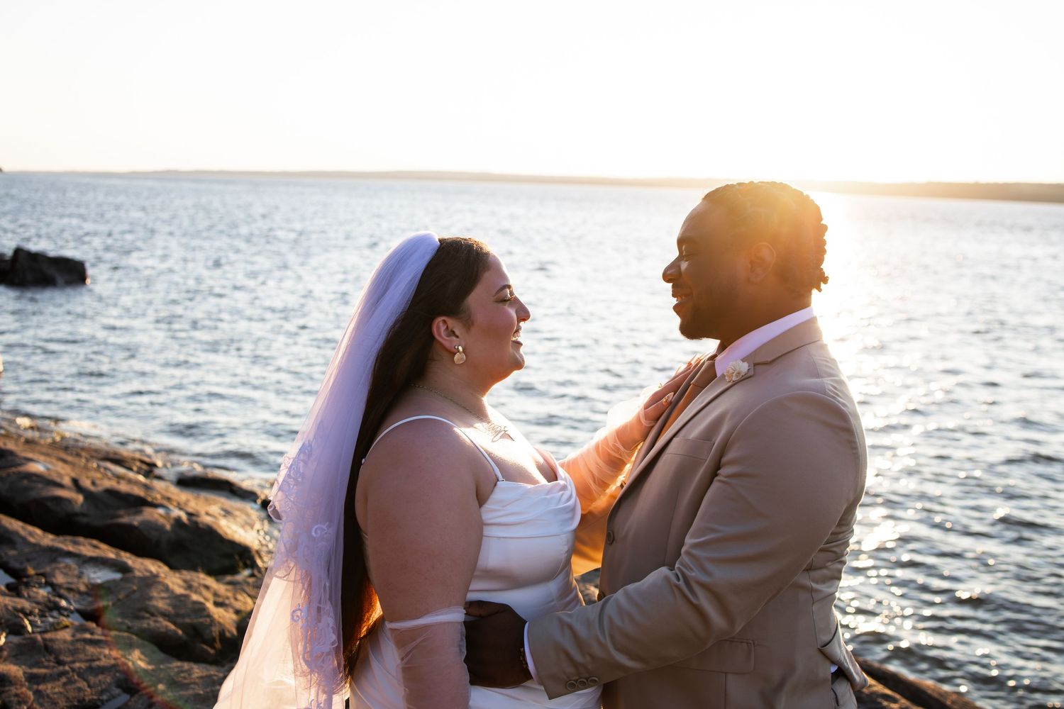 Interracial couple sharing a romantic moment by the ocean at sunset, bride in white gown and veil, groom in tan suit.