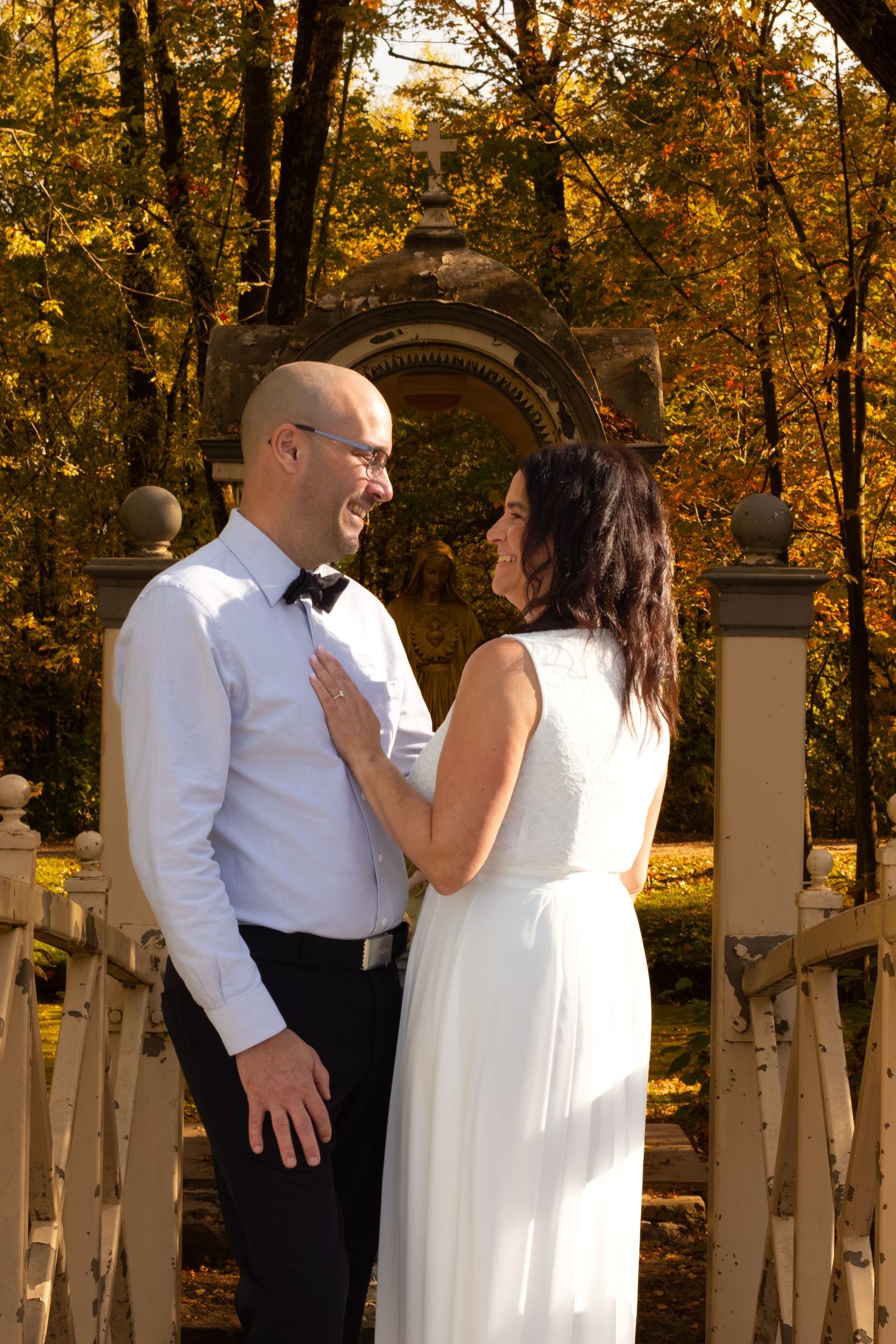 Smiling bride and bald groom in bow tie sharing a romantic moment on a bridge in autumn park.