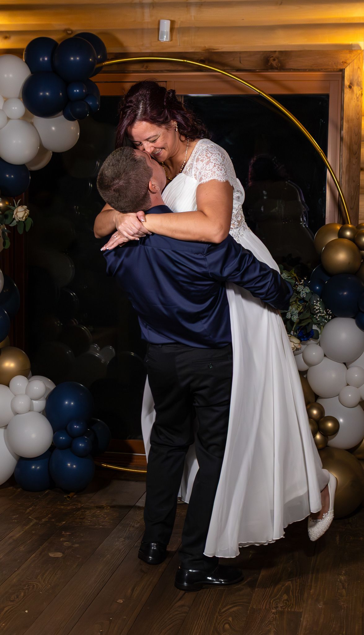 Groom lifts bride in white lace dress in front of elegant navy and gold balloon arch at wedding celebration.