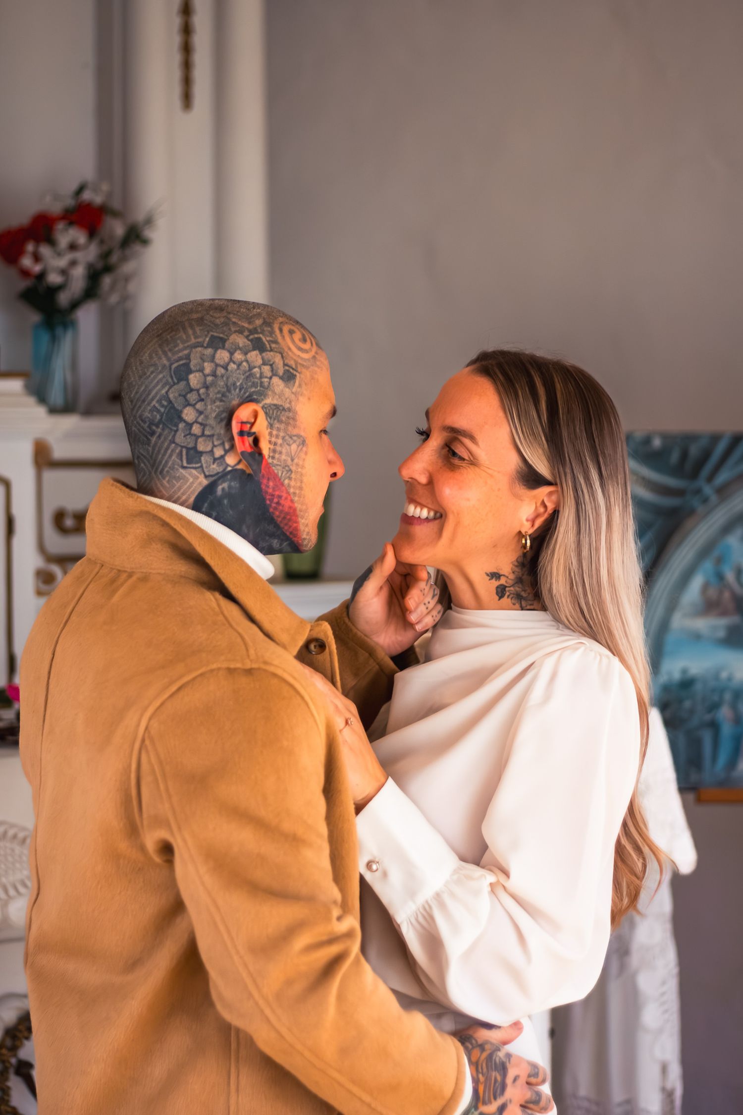 Smiling couple embracing, man with full head tattoos wearing brown jacket, woman in white blouse touching his face.