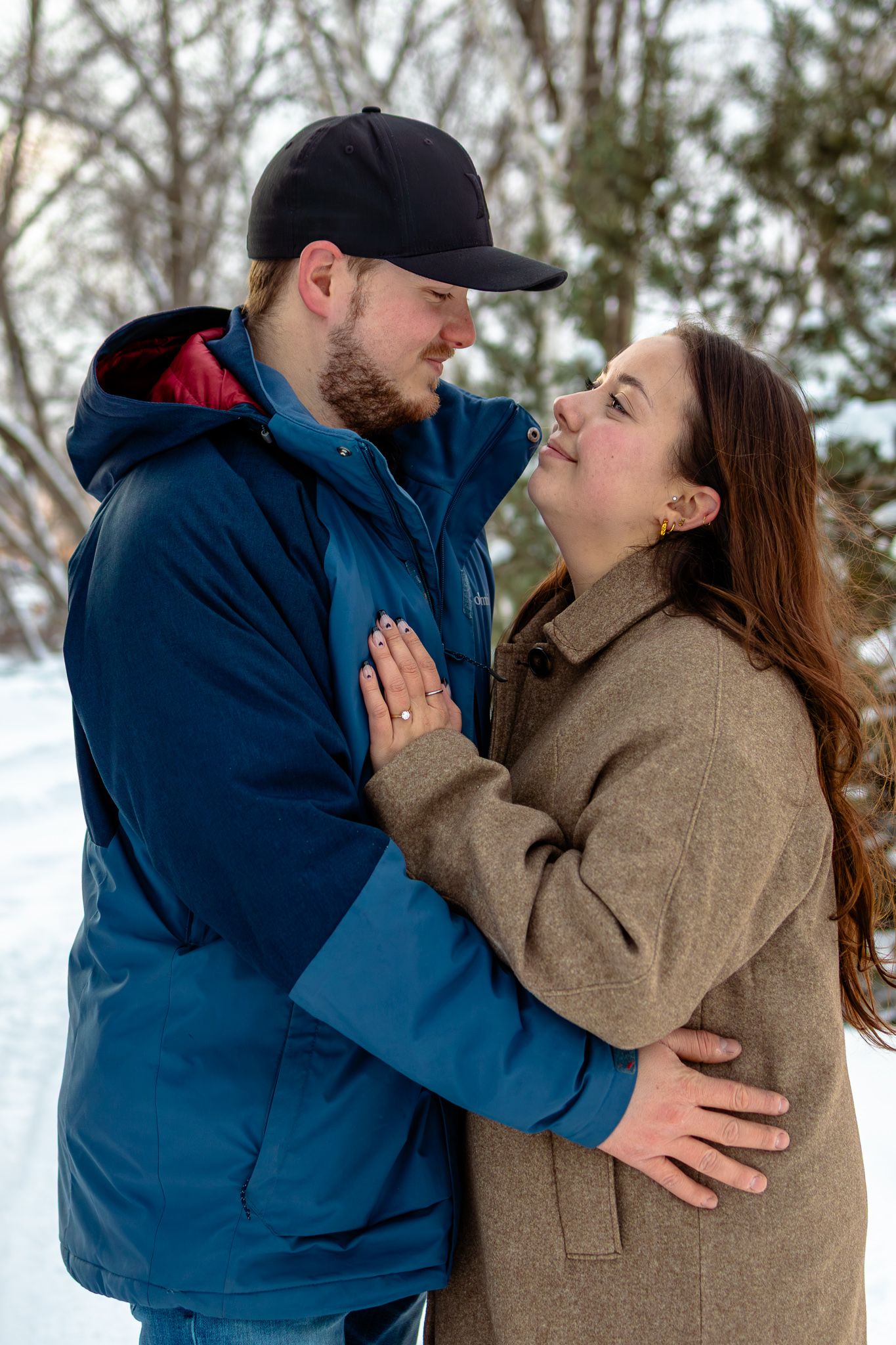 Couple embracing and gazing into each others eyes in snowy winter setting, woman displaying diamond engagement ring on her hand.