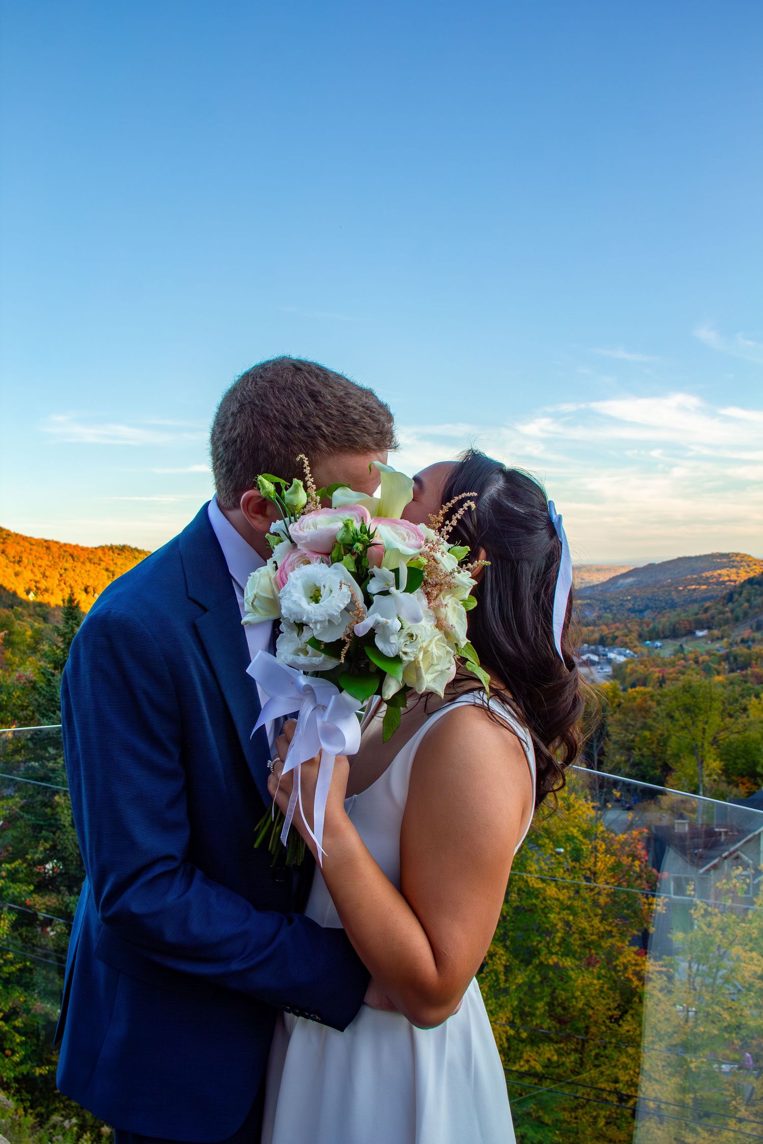 Newlyweds kiss behind a white floral bouquet with colorful autumn foliage and blue sky in the background.