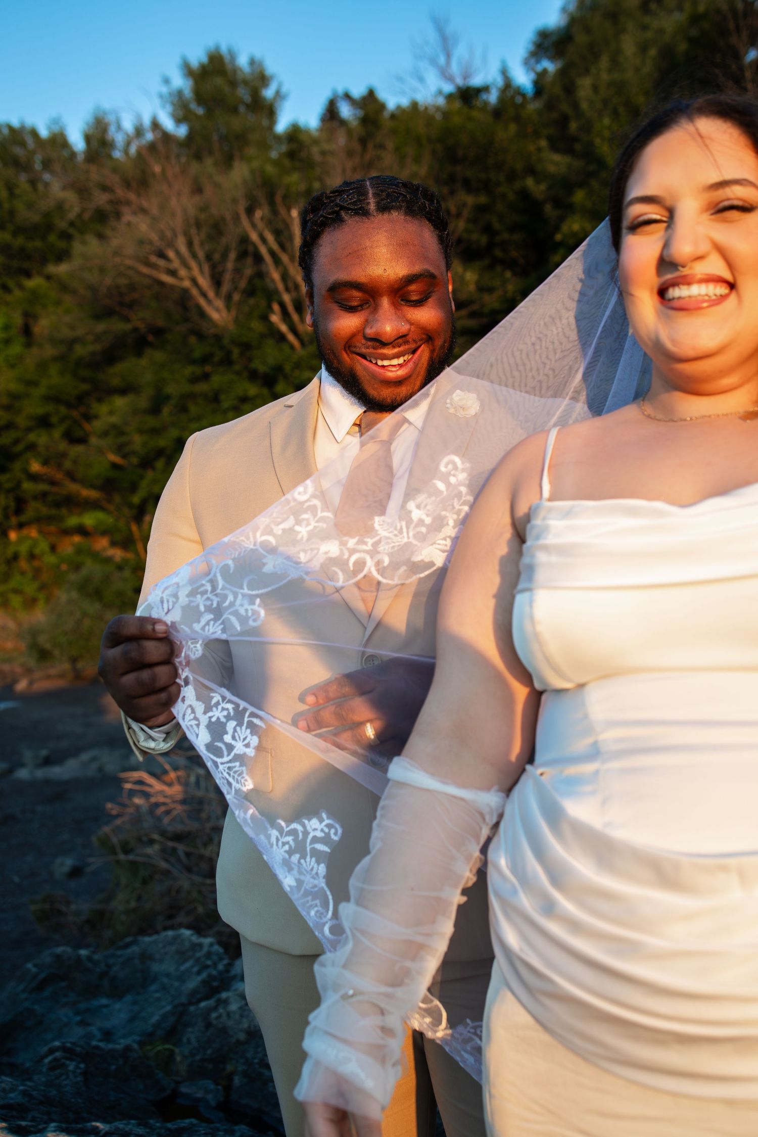 Smiling interracial couple in wedding attire outdoors, groom in tan suit holding bride's lace veil at golden hour.