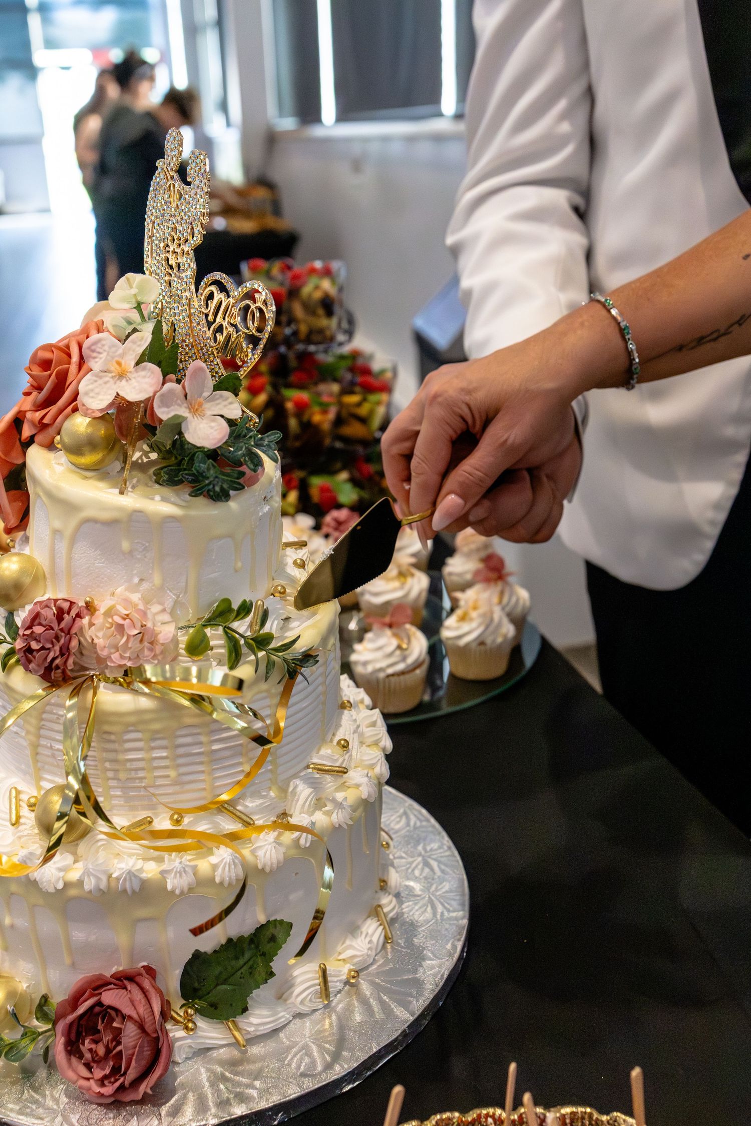 Person cutting a elegant three-tier white wedding cake decorated with roses, gold accents, and floral topper at a reception.