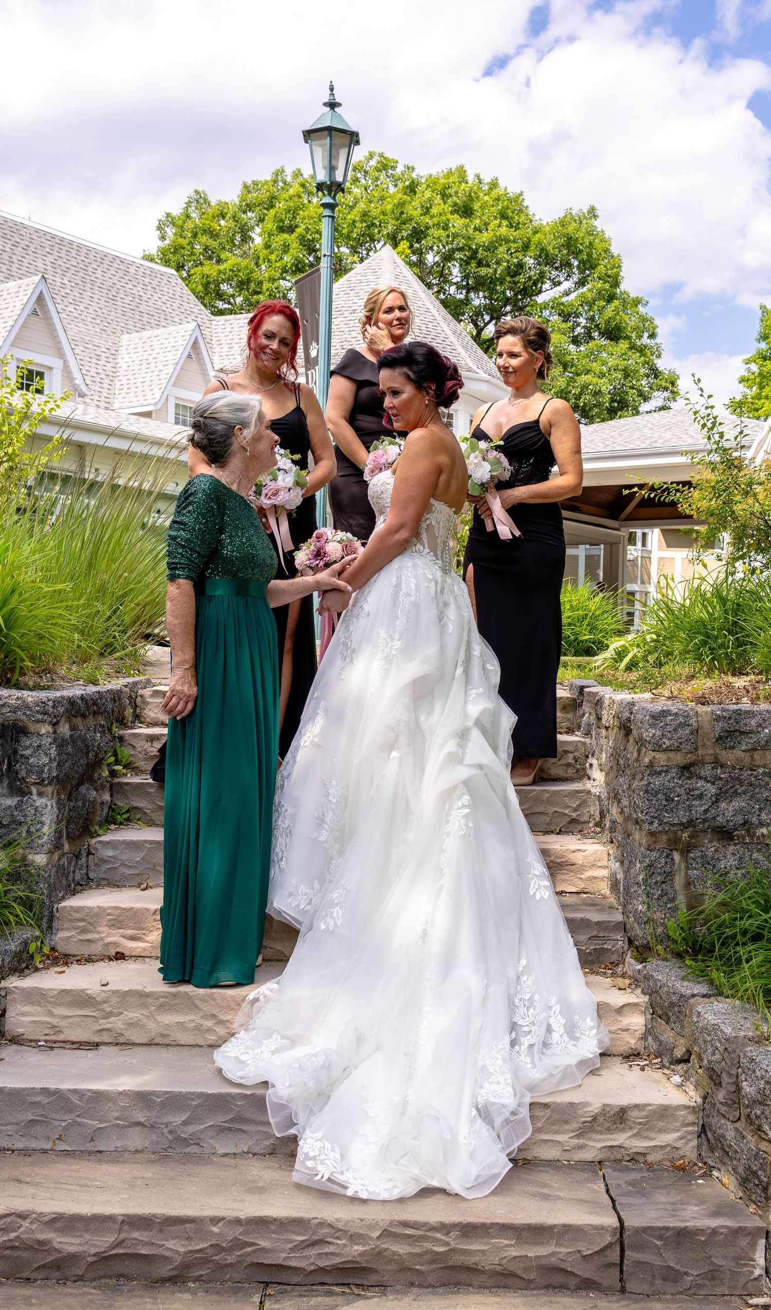 Bride in white lace gown stands on stone steps with bridesmaids in black and green dresses, holding bouquet outdoors.