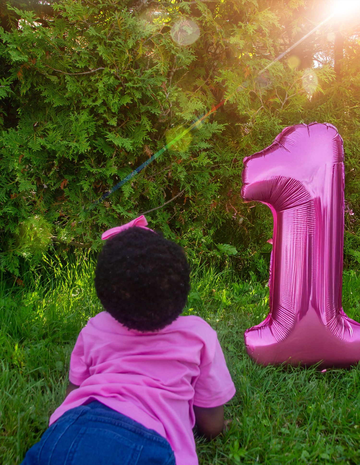 Toddler in pink shirt lying on grass beside a large pink number 1 balloon on their first birthday.