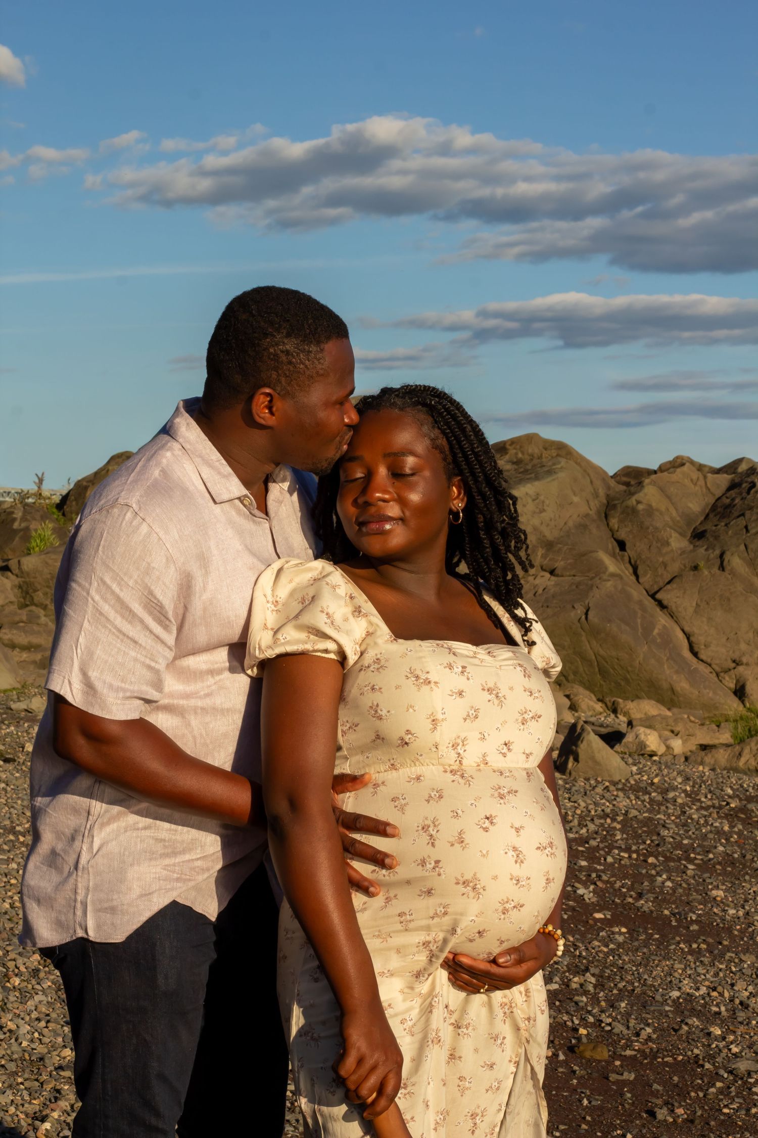 A man kisses a smiling pregnant Black woman in a floral dress on a rocky beach at sunset.