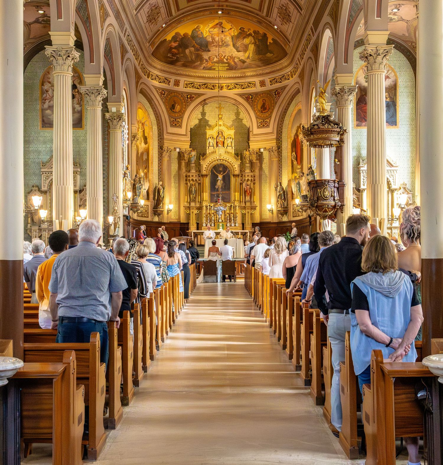 Congregation gathered inside ornate Catholic church with wooden pews, gilded altar, and painted ceiling during Mass.