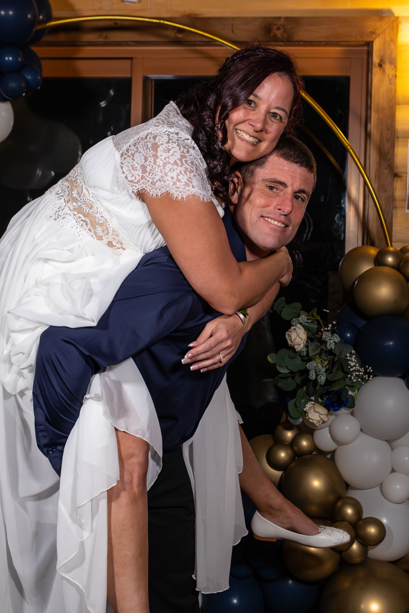 Groom giving bride a piggyback ride in front of navy blue and gold balloon arch at wedding reception.