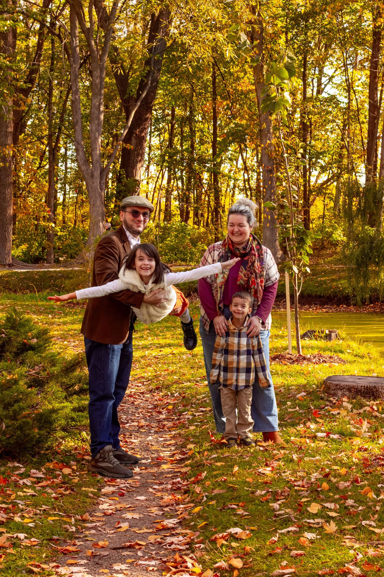 A joyful family of four plays in an autumn park, grandfather swinging granddaughter on a leaf-covered path.