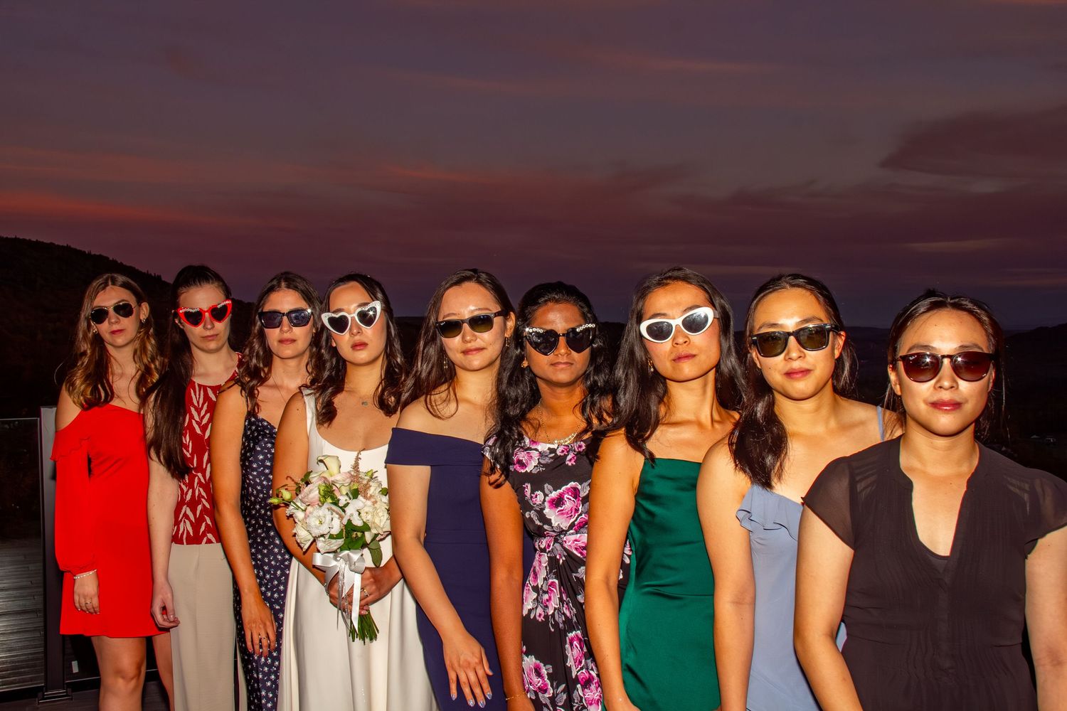 Bride and bridesmaids wearing matching white sunglasses pose together in colorful dresses at a sunset wedding.