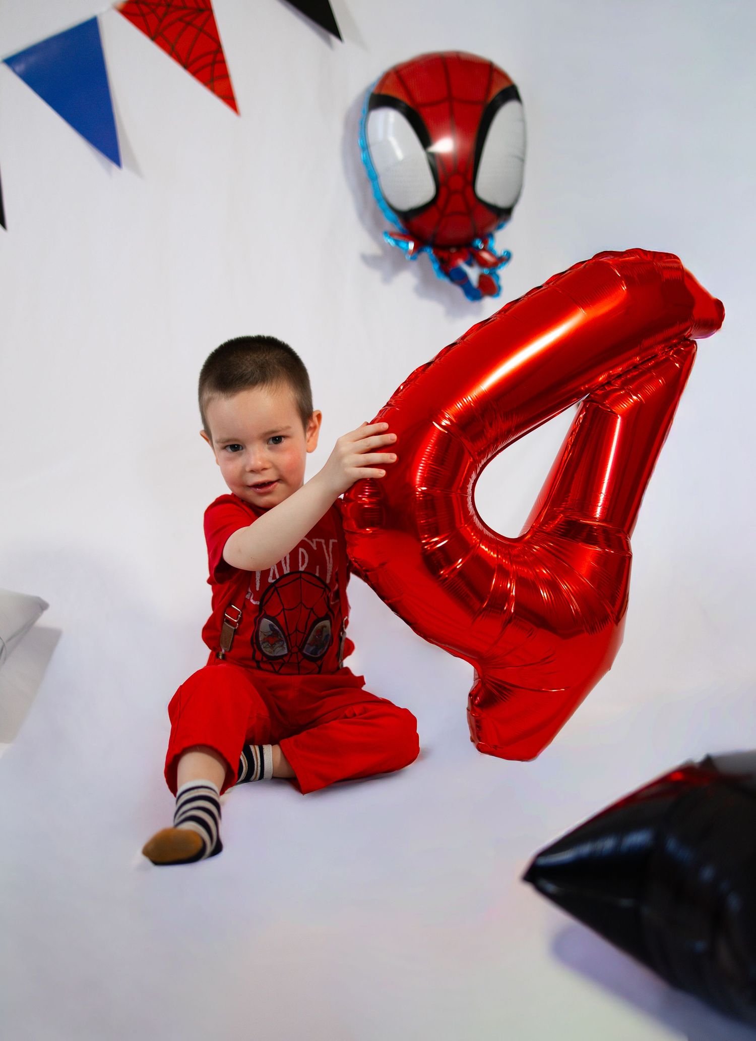 A toddler in red Spiderman outfit sits beside a large red number 4 balloon at a Spiderman themed 4th birthday party.
