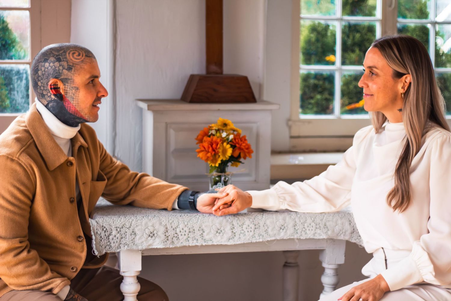 Couple sitting across from each other at a lace-covered table holding hands in a bright room.