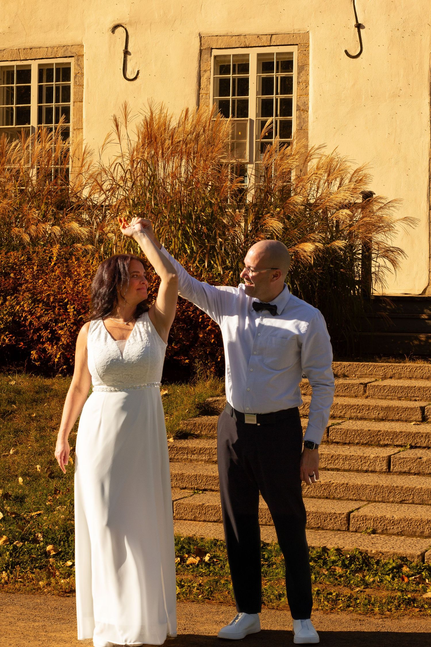 Bride and groom dancing outdoors in autumn sunlight near a historic building.